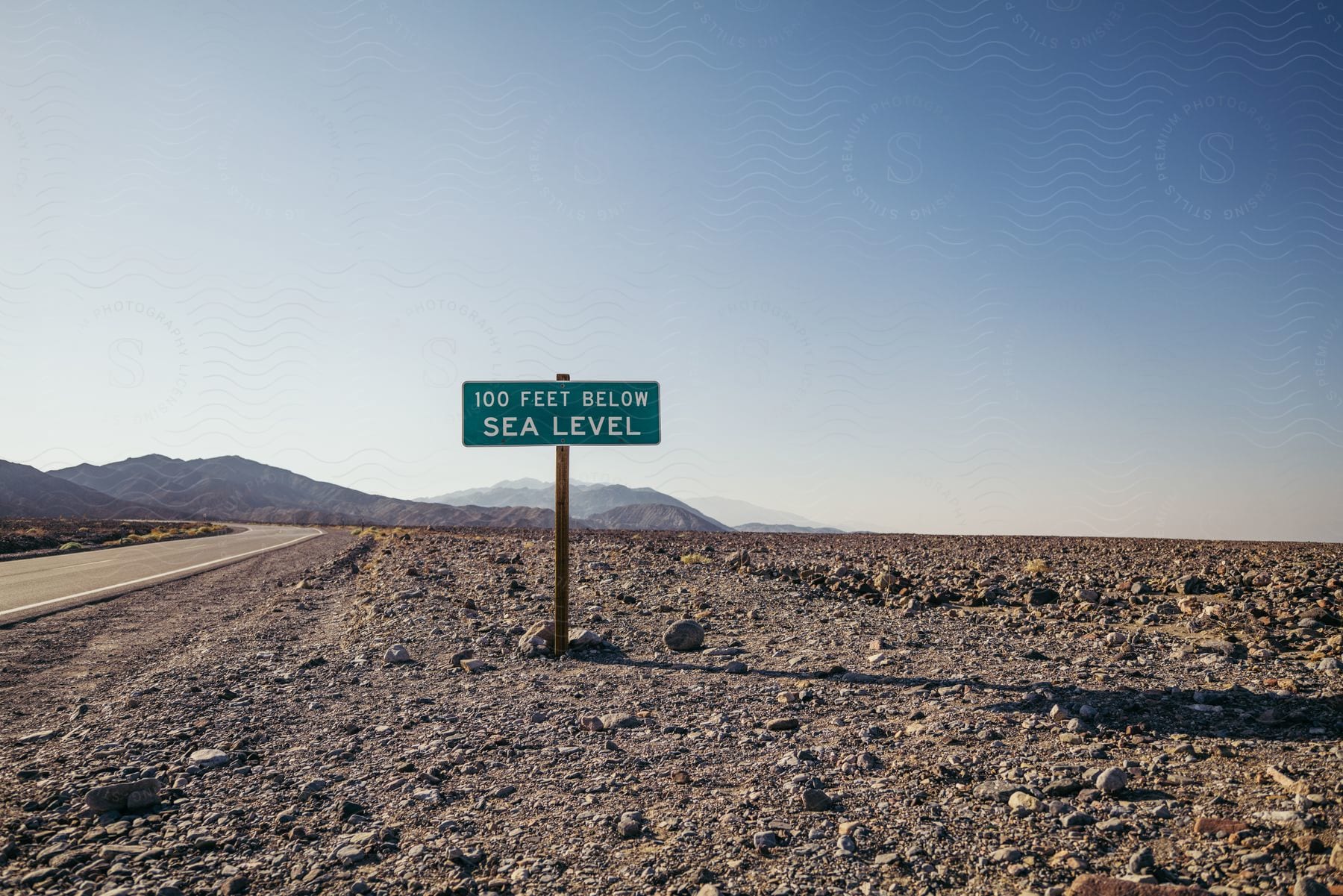 Roadside sign stating 100 feet below sea level and mountains in the distance under a clear sky