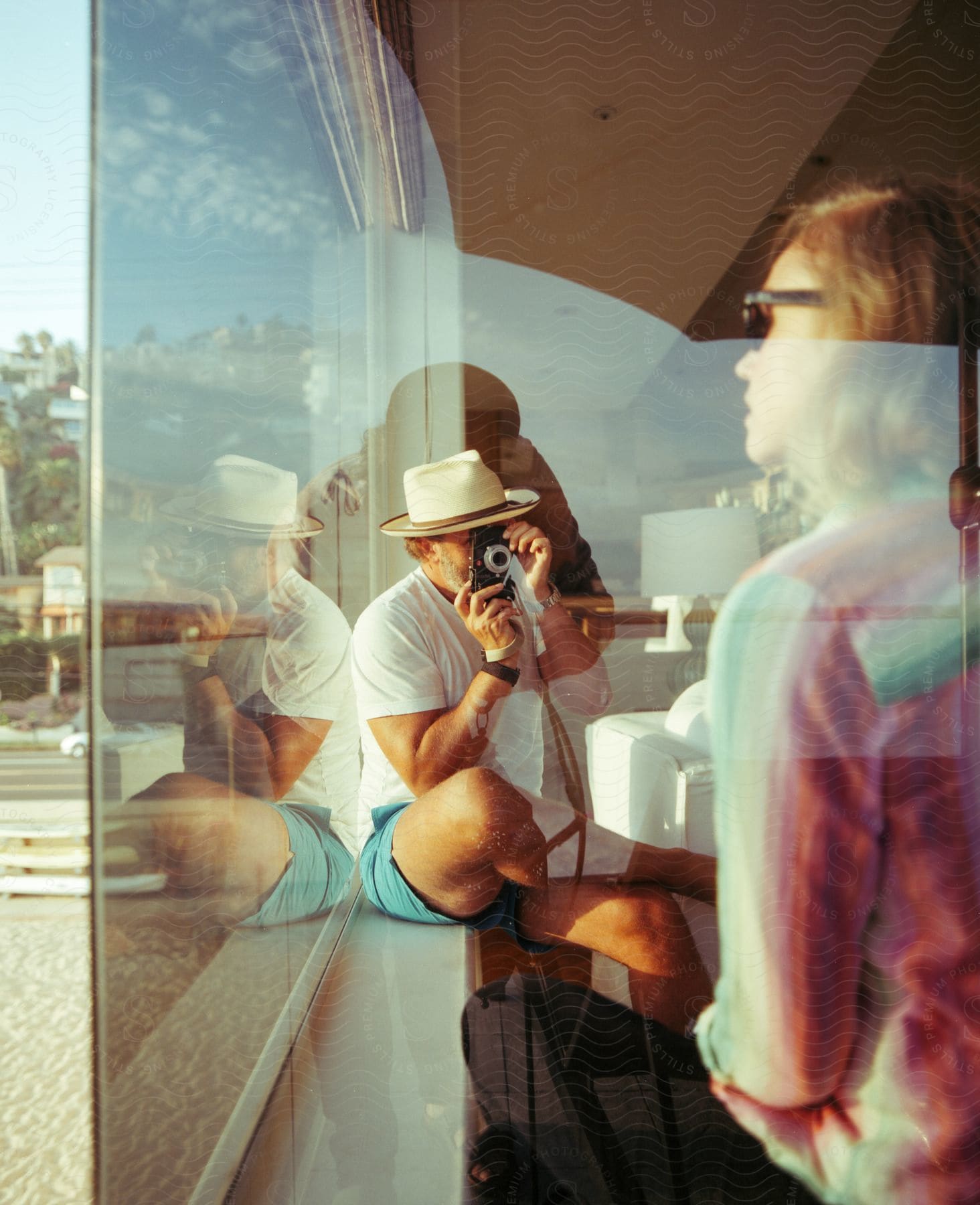 A man sitting on a window sill takes a photo of a woman looking out of a window