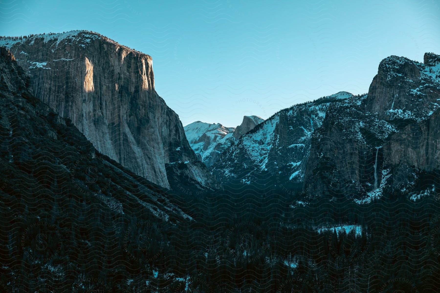 A Mountain Cliff Overlooks A Forested Valley With Mountains In The Distance Under A Clear Blue Sky