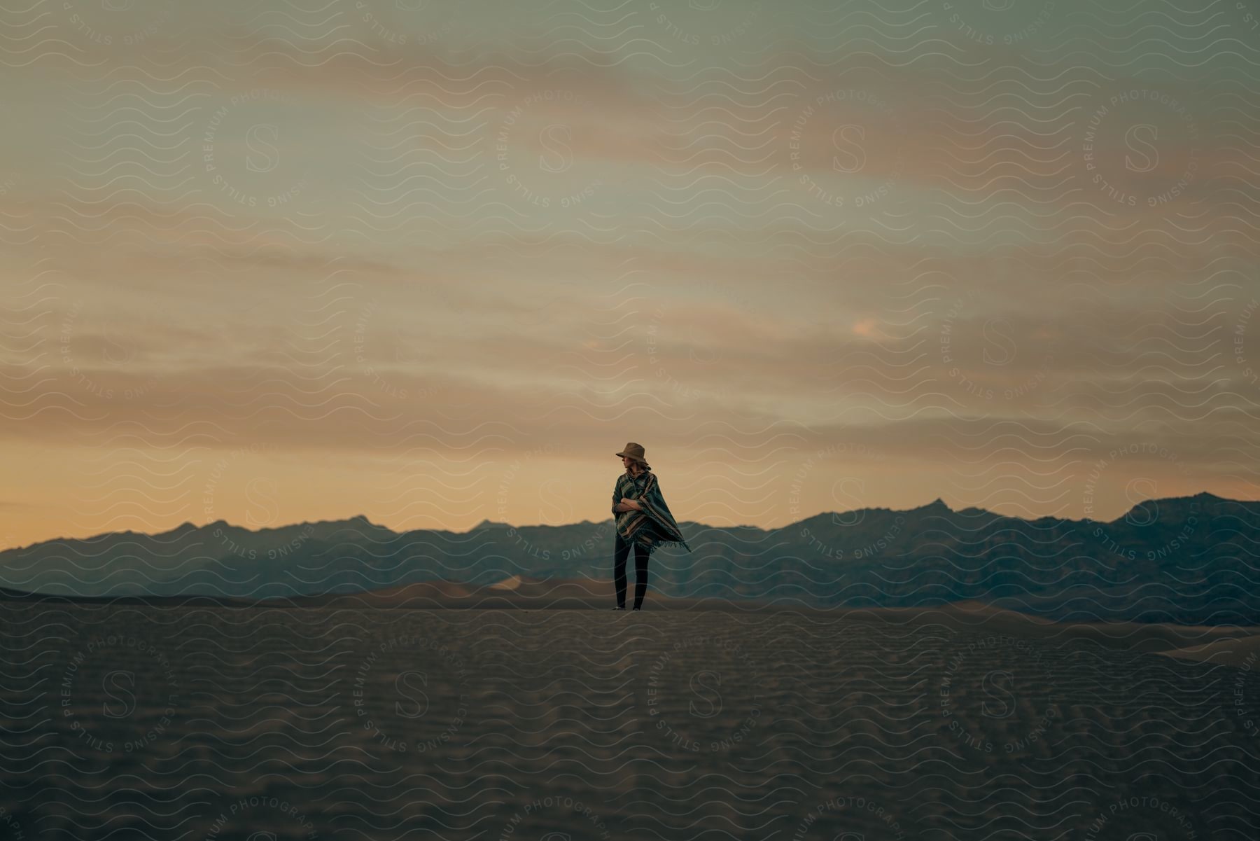 Stock photo of woman wearing a poncho and hat standing in a desert before mountains at sunset