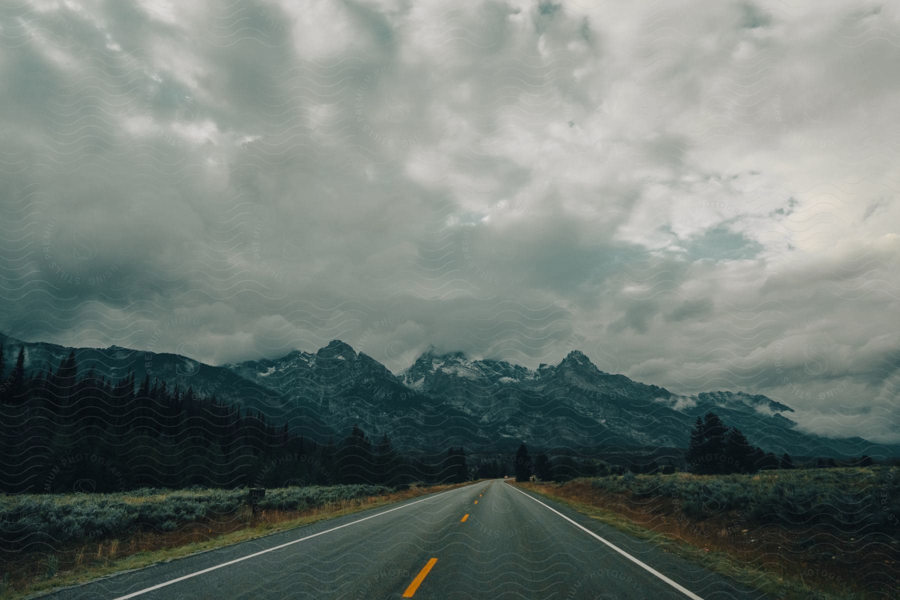 An open road leads to snow covered mountains and a forest under a cloudy sky in the outdoors during winter