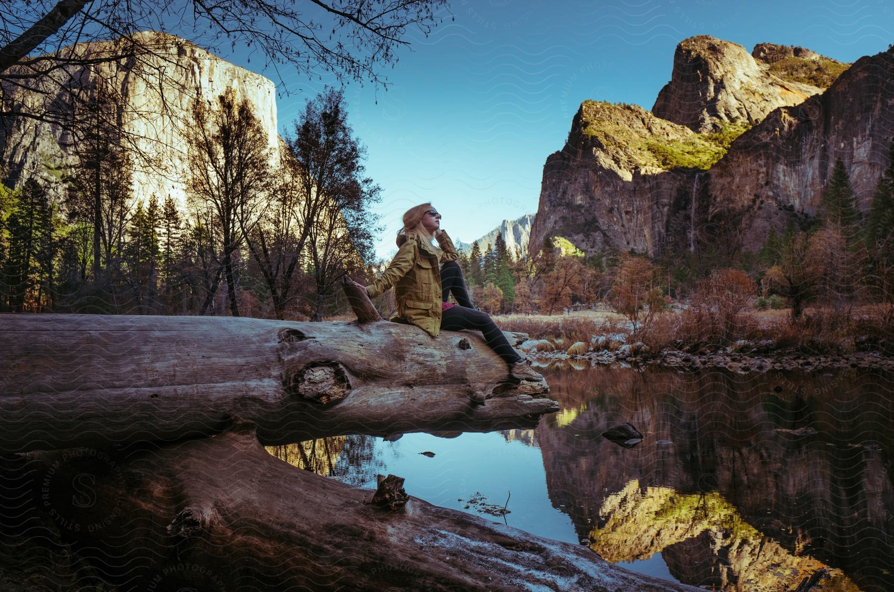 Blonde woman in sunglasses sits on fallen tree over serene lake watching mountains in the background