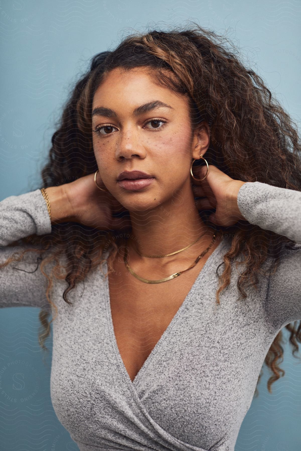 A young woman with long wavy brown hair wears a grey wrap dress hoop earrings gold necklaces bracelet and poses with her arms raised behind her head in front of a baby blue background