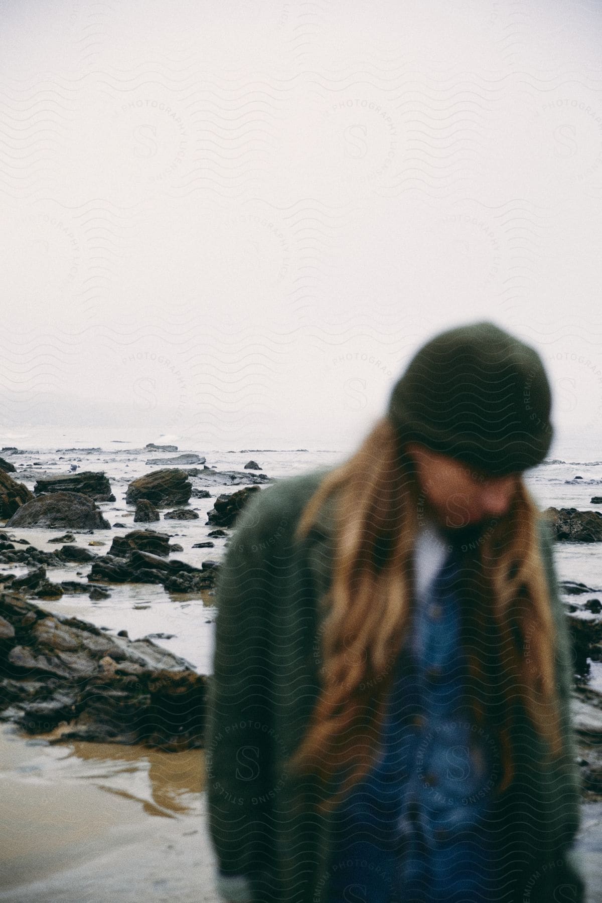 A person wearing a coat and hat stands on a rocky shoreline by the sea