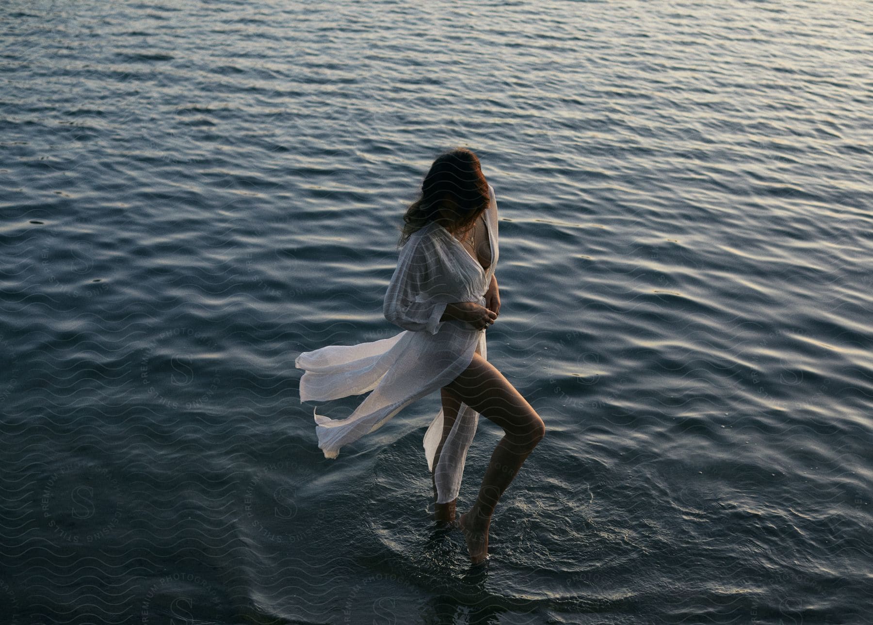 A woman standing in shallow beach waters surrounded by serene outdoor scenery at dusk wearing a flowing white gown