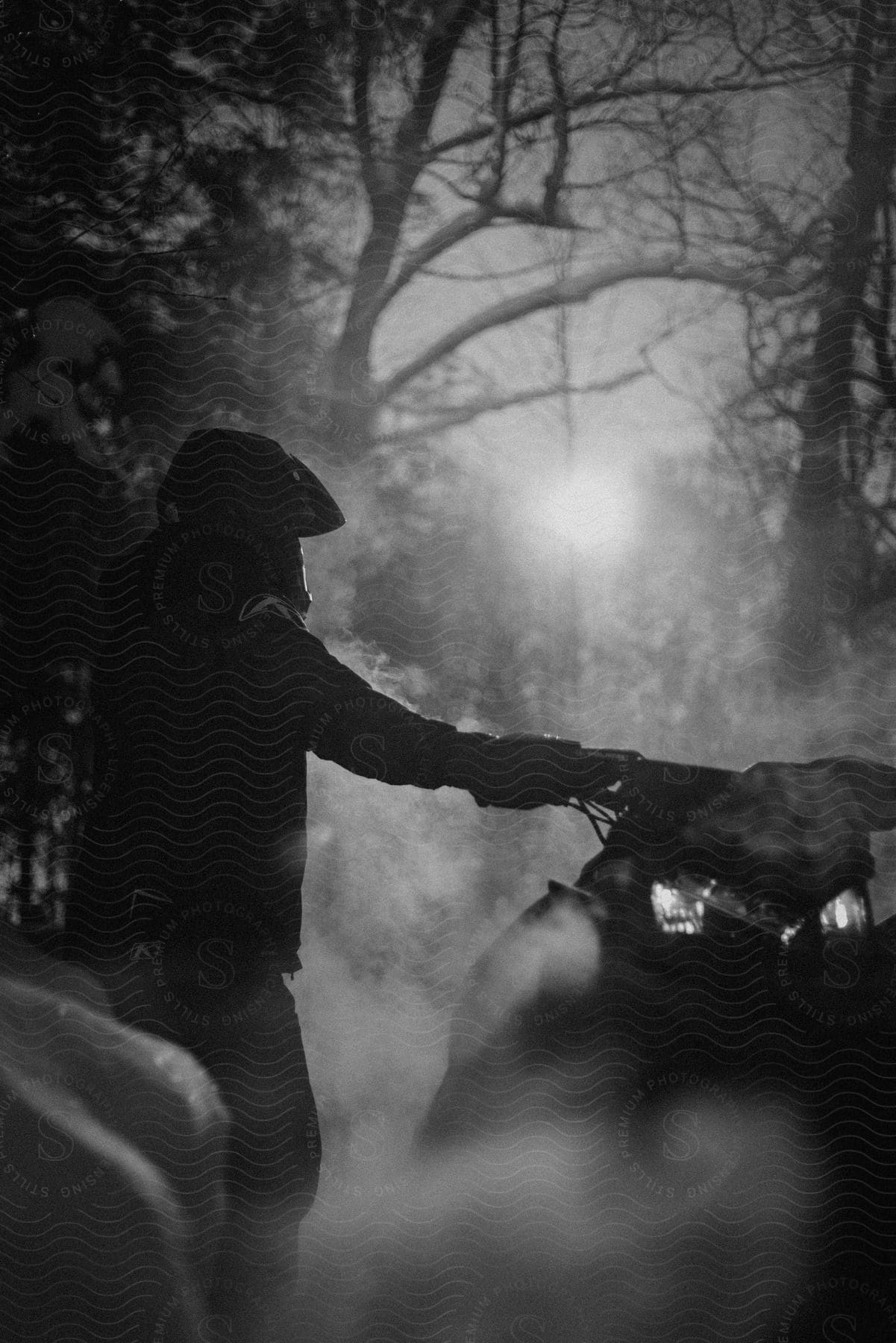 A man in riding gear and a helmet stands next to an atv in the moonlit foggy woods