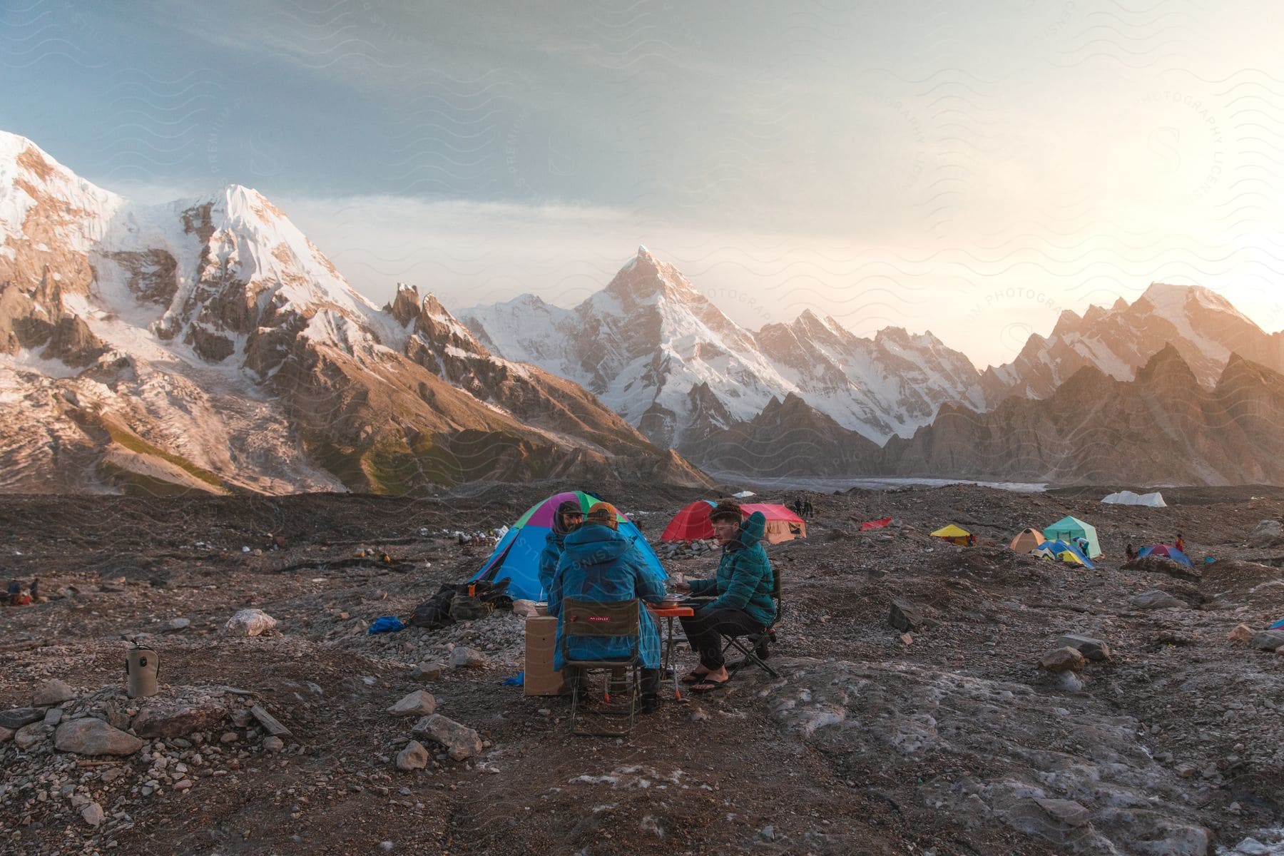 A small group of hikers with tents on a rocky terrain at dusk near snowy mountains