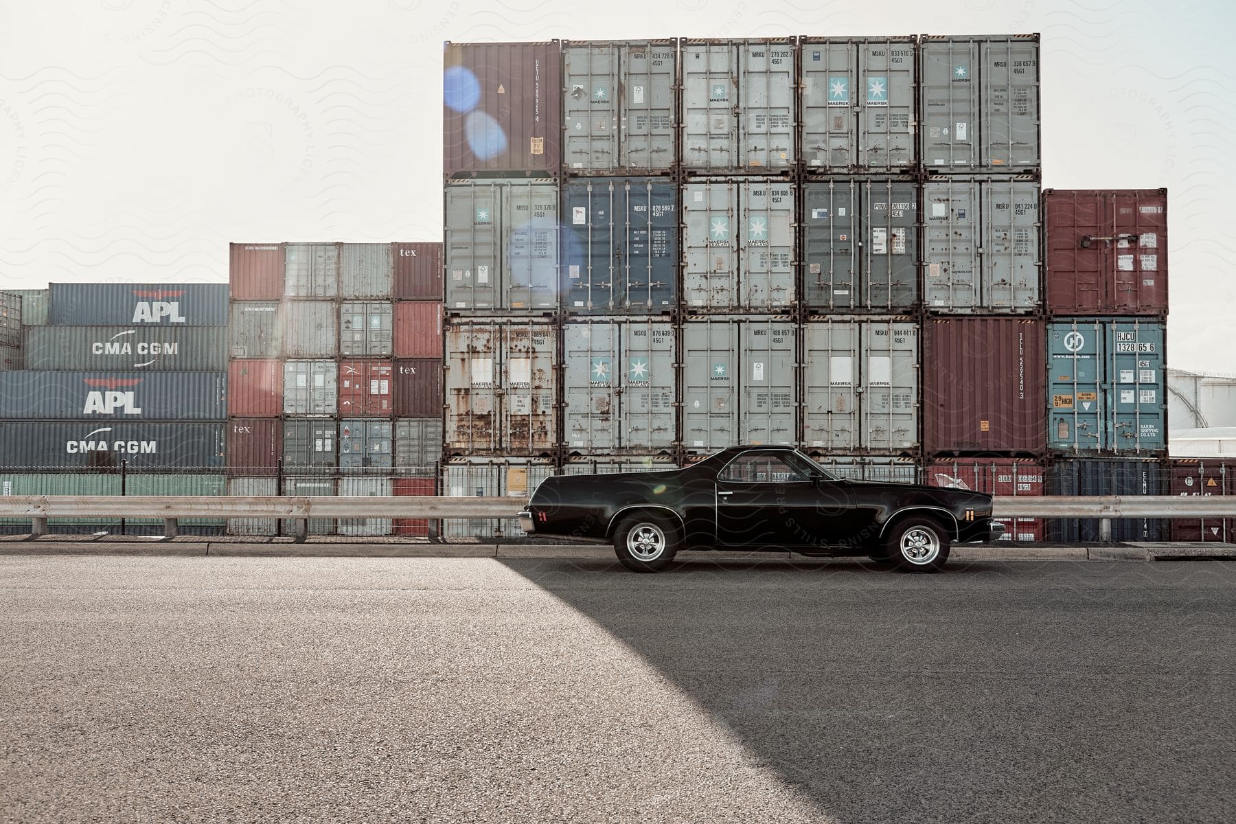 A black el camino is parked near a guardrail and stacks of shipping crates