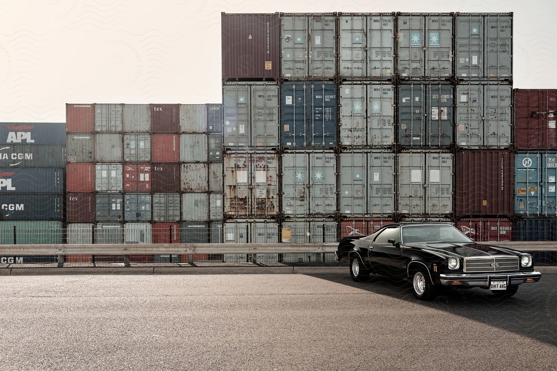 A black el camino parked near a guardrail and stacks of shipping crates