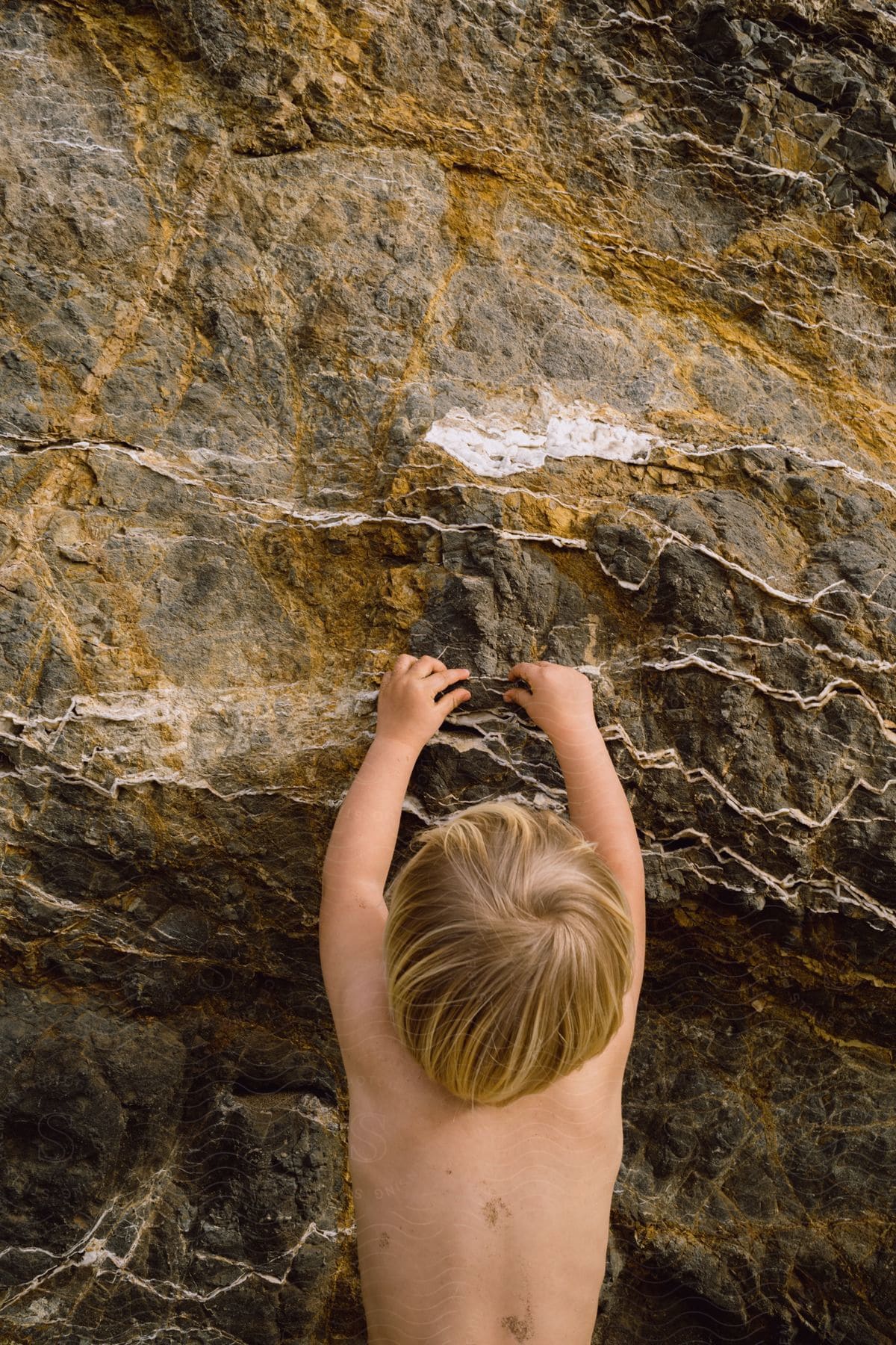 Blonde toddler stretches arms to touch zigzag rock layers at a cliff face