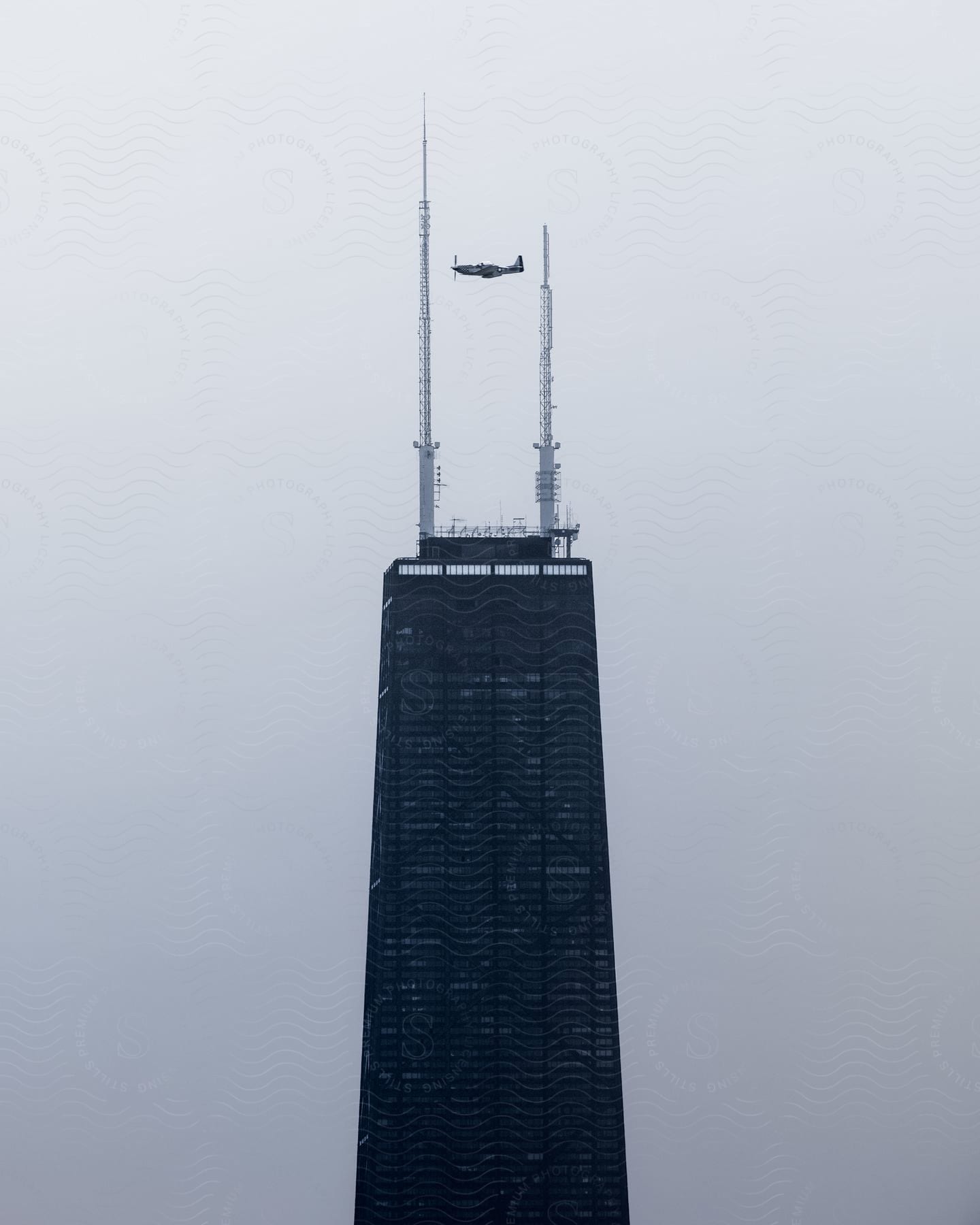 A tall skyscraper building with two poles on the top as a plane flies over the building in chicago