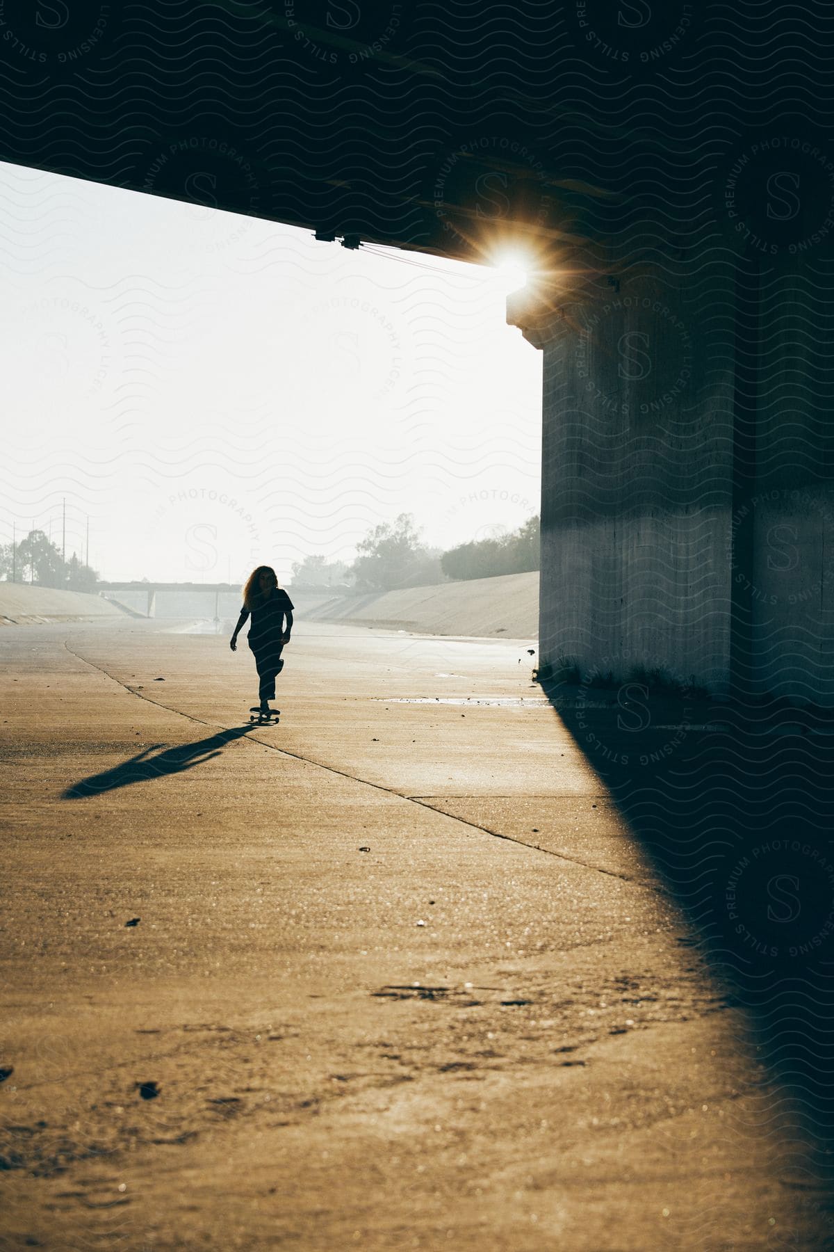 Woman skateboarding under bridge is silhouetted against the sun