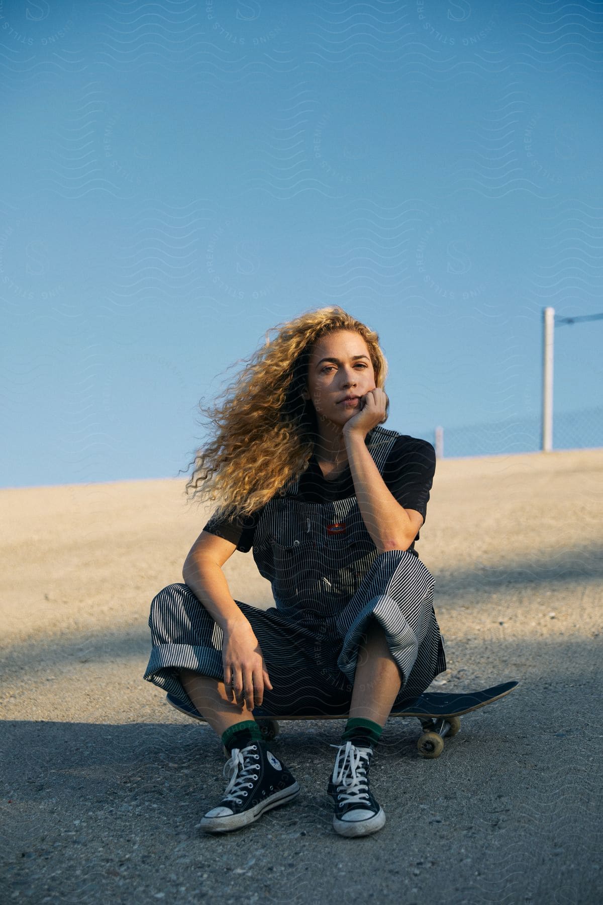 Teen girl skateboarder sits on skateboard chin on hand windblown curly hair black tshirt pinstripe overalls black converse hightops