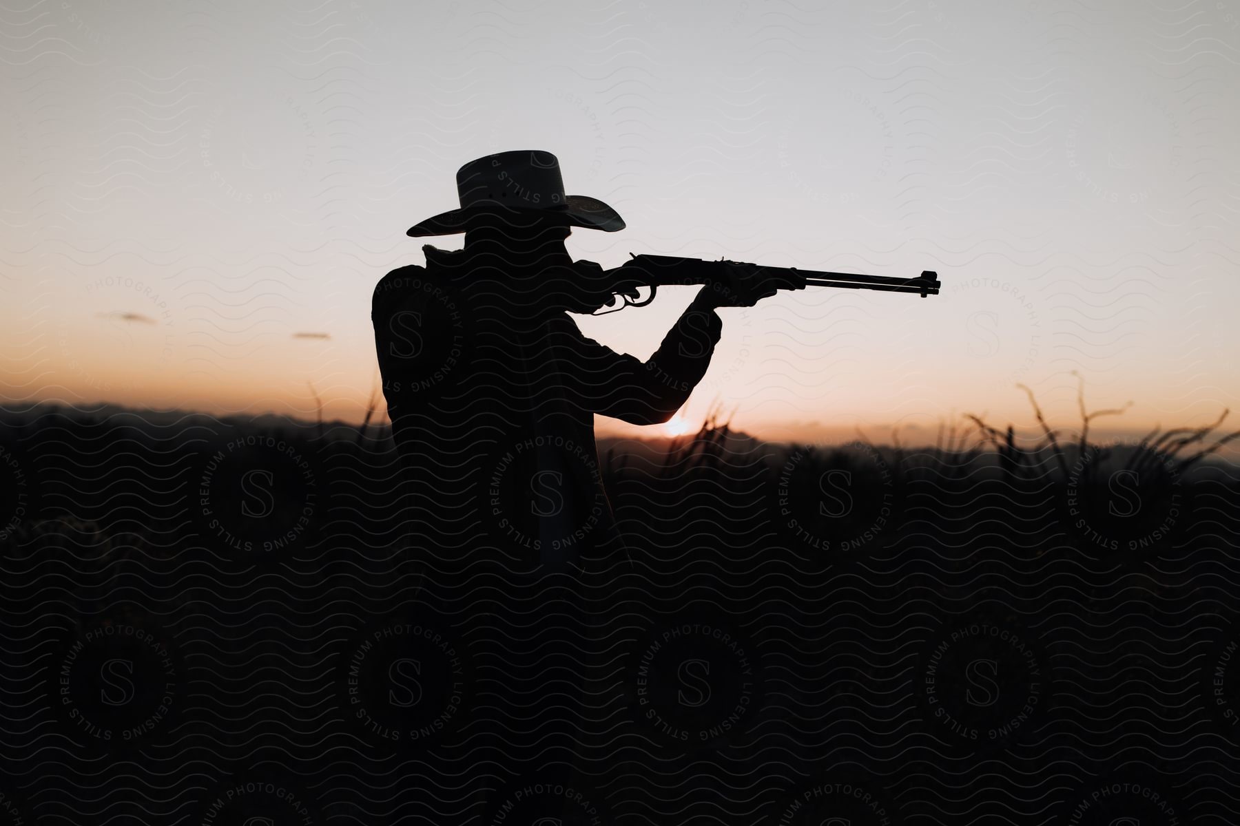 Silhouette of a man wearing a cowboy hat aiming a rifle with mountains in the distance