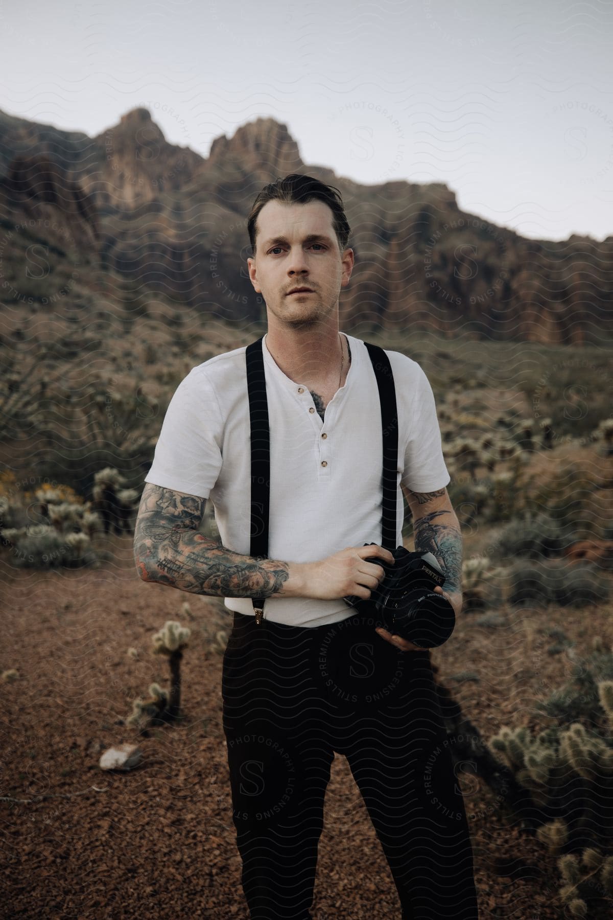 A man stands in the desert holding a camera with mountains and rock formations in the distance