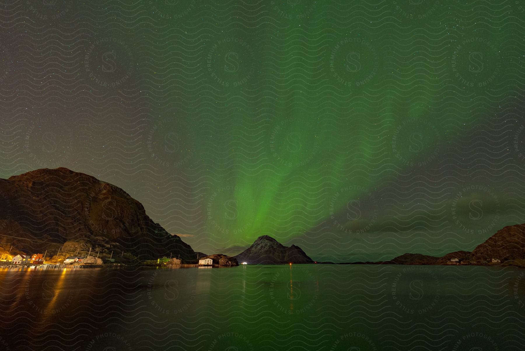 Aurora australis dances above a serene lake nestled among majestic mountains in a coastal town at night