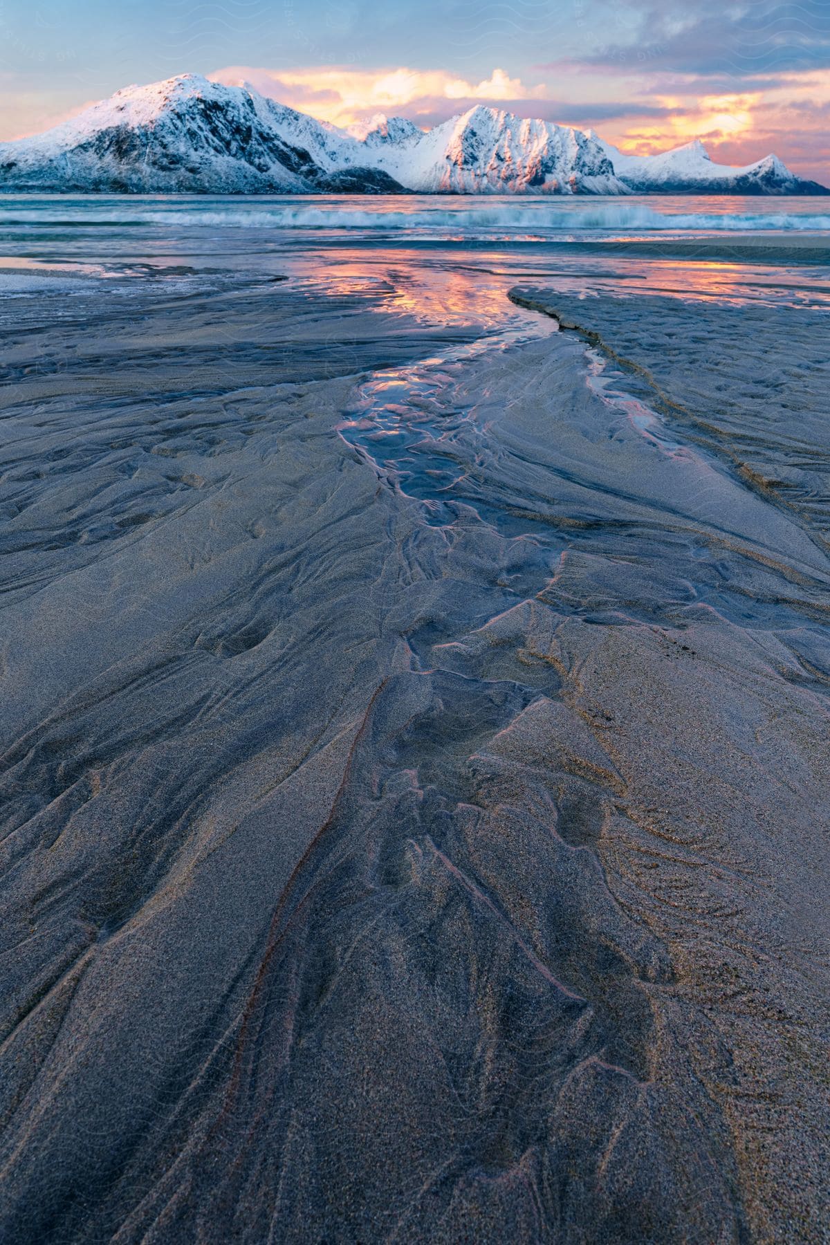 A snowy mountain reflects vibrant colors at sunset over a sandy shore