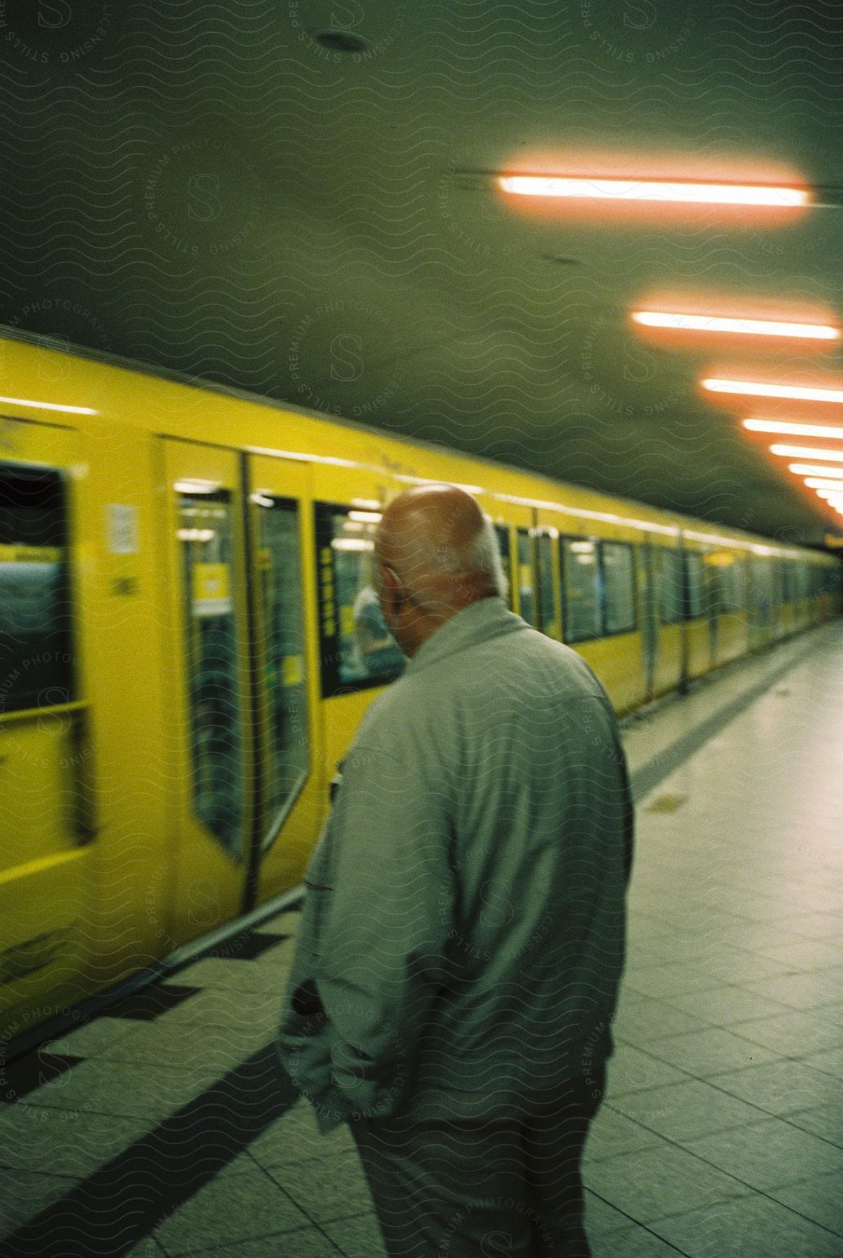An older man waits alone on a concrete platform as a yellow subway train arrives