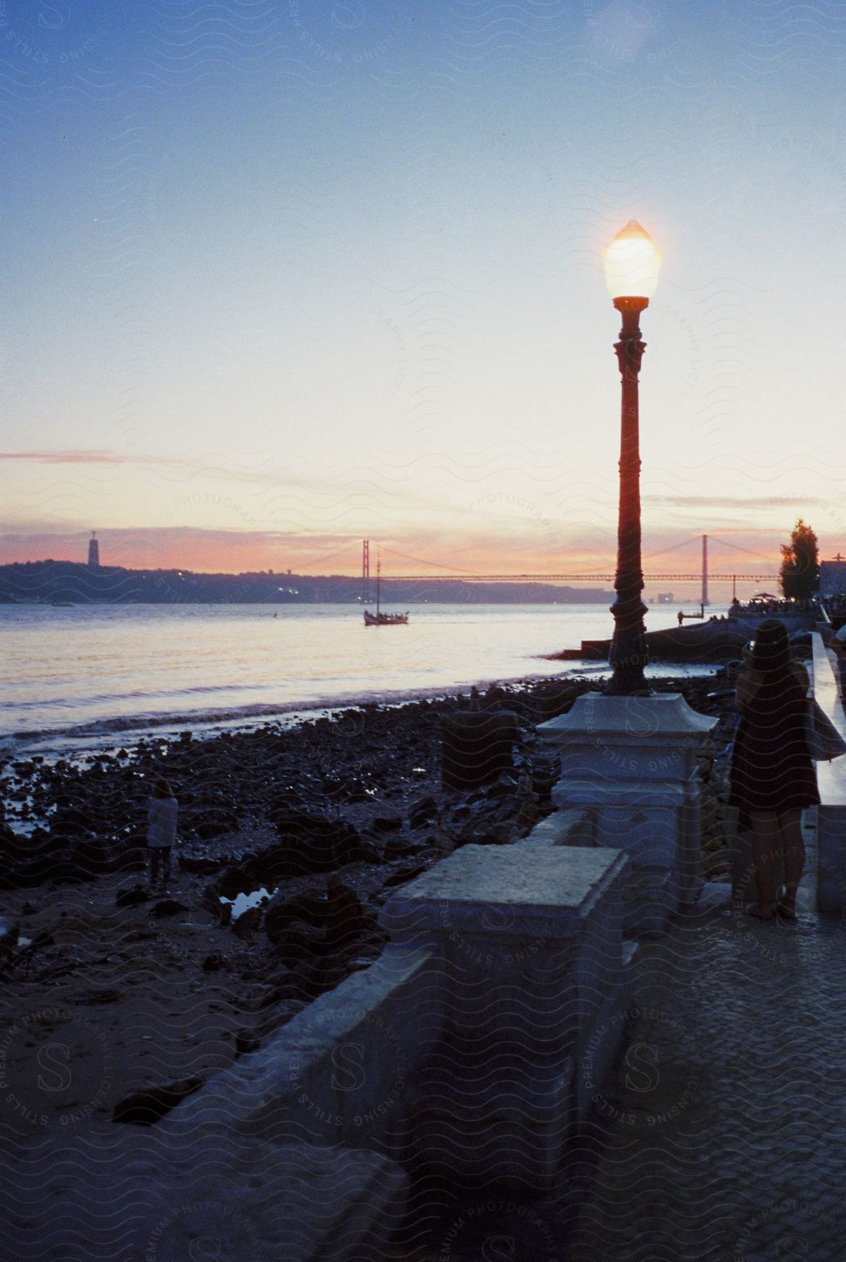 A woman is photographing another person by the sea with a bridge on the horizon