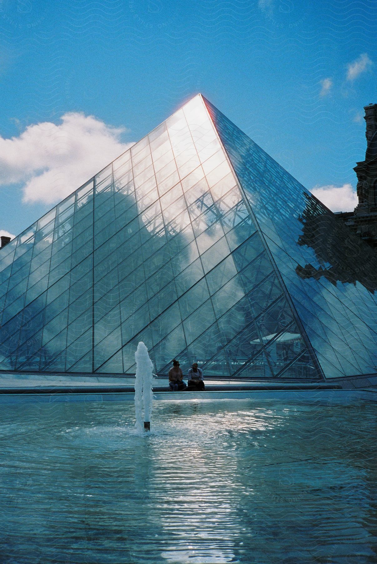 Two people sit on a bench in front of the louvre with a fountain on a cloudy day