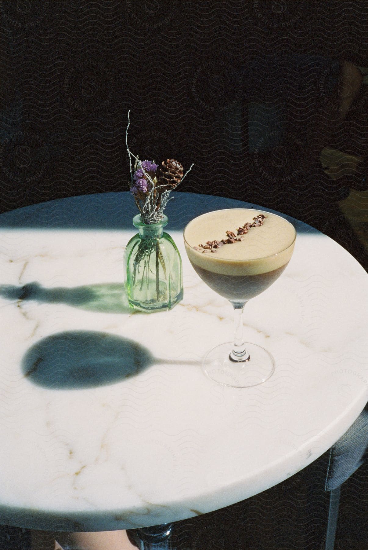 A coffee cocktail on a marble bistro table next to a glass bud vase with dried flowers late afternoon