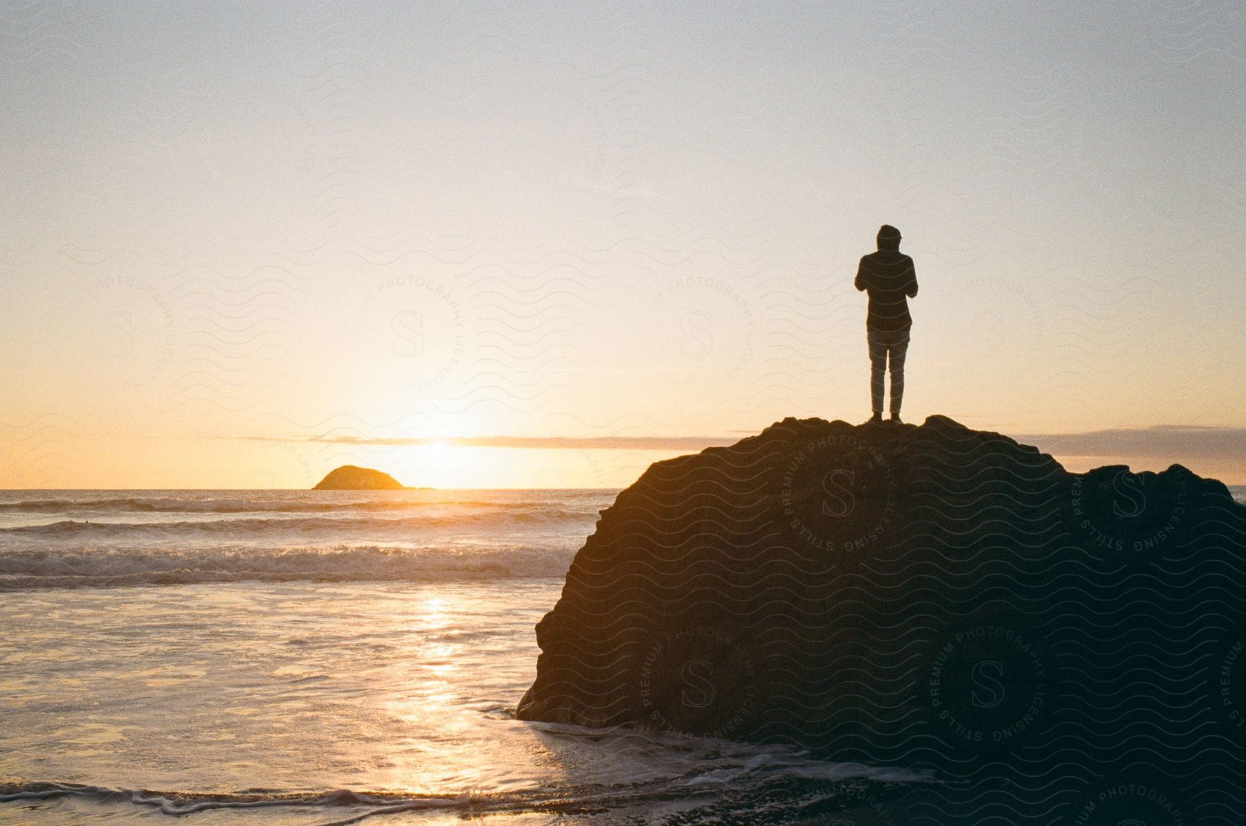 Person standing on rock as waves roll in and sun sets on horizon