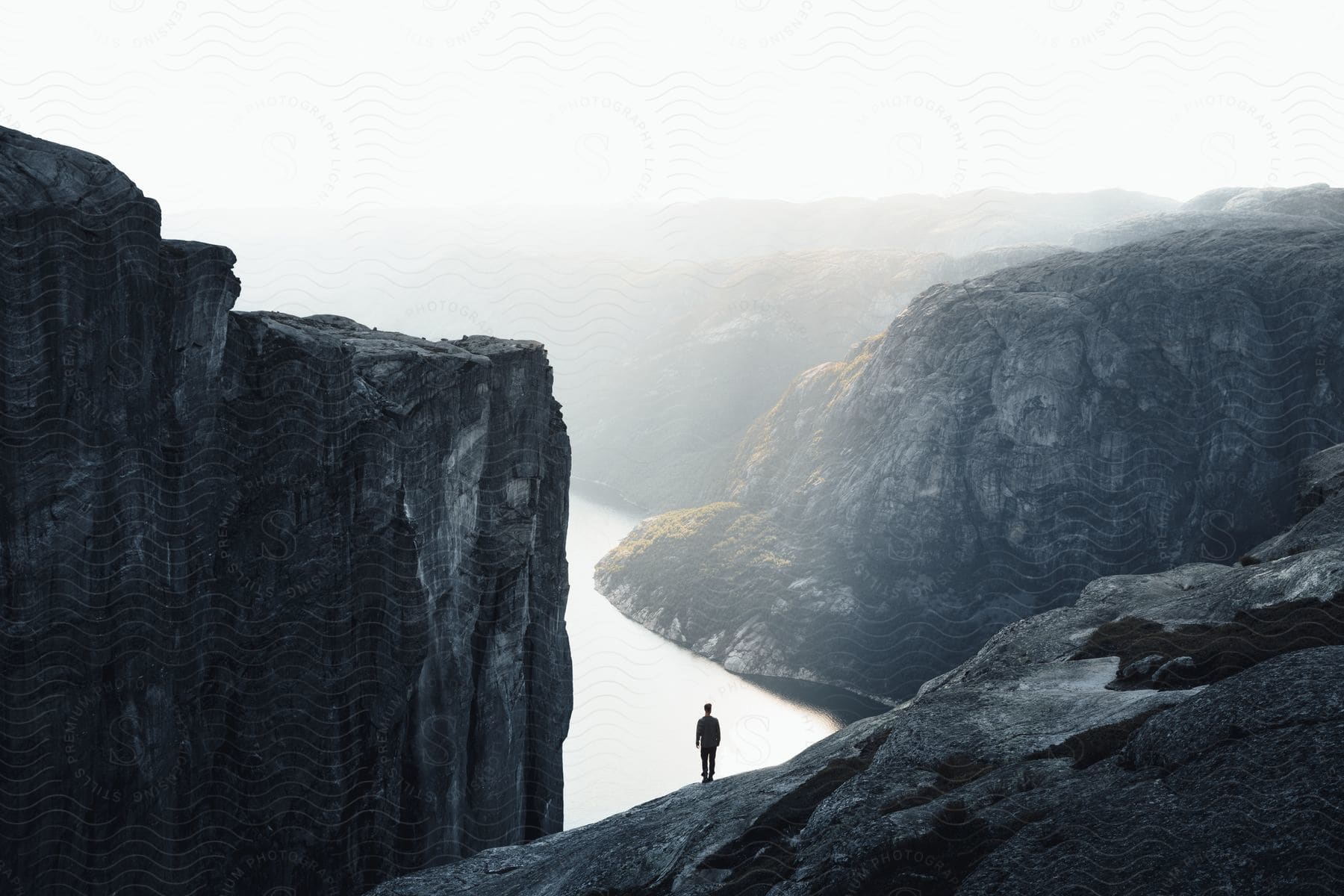 A man standing on a mountain slope overlooking a river valley under a foggy sky