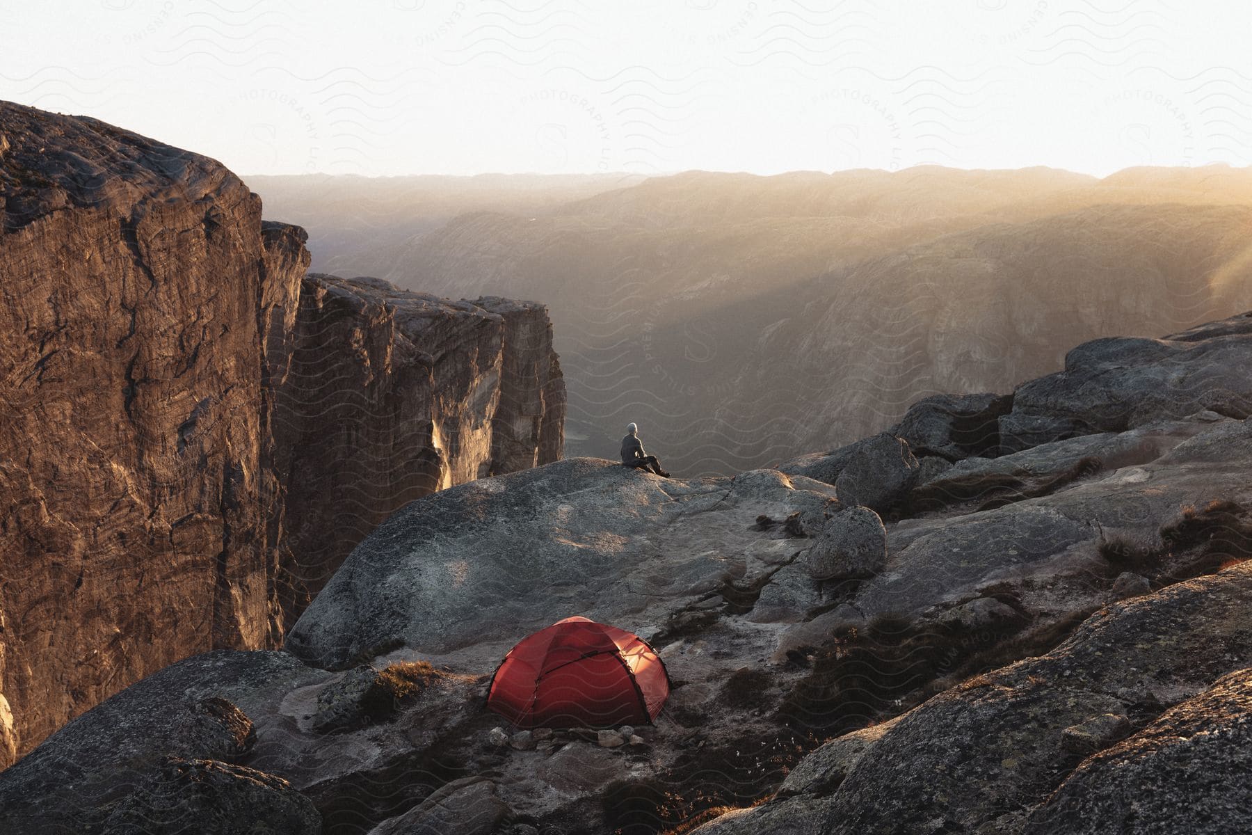 A person sits on a ledge near a red tent overlooking a canyon as the sun shines over mountains in norway