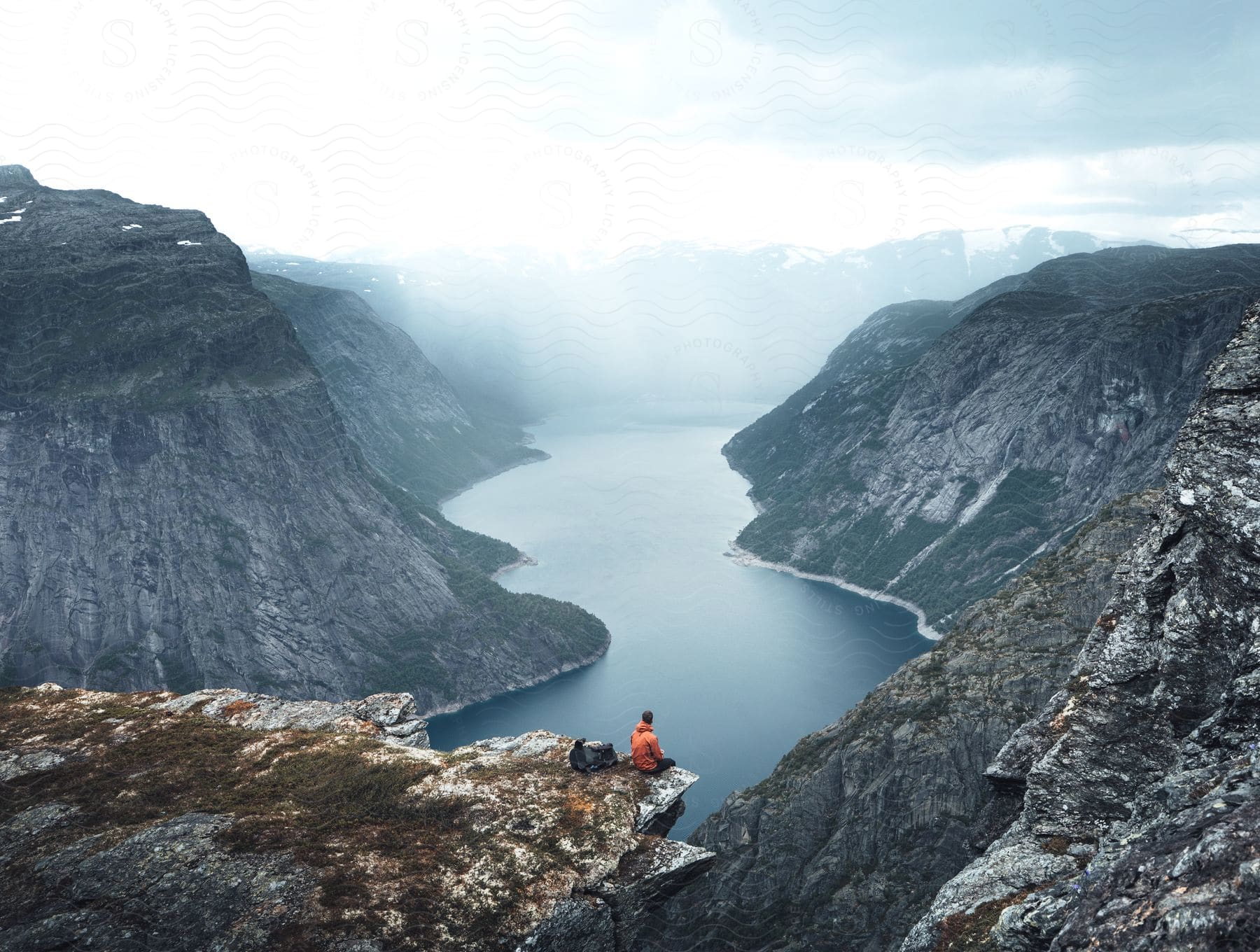 Stock photo of man in orange jacket sitting on mountain edge looking at mountain range and wide river