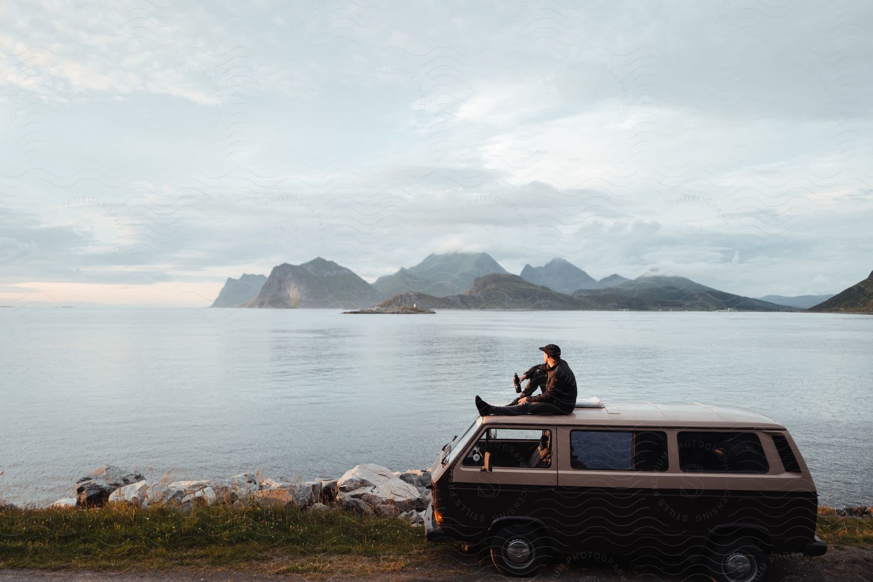 A man sits on his van as he looks out over the sea toward distant rocky cliffs