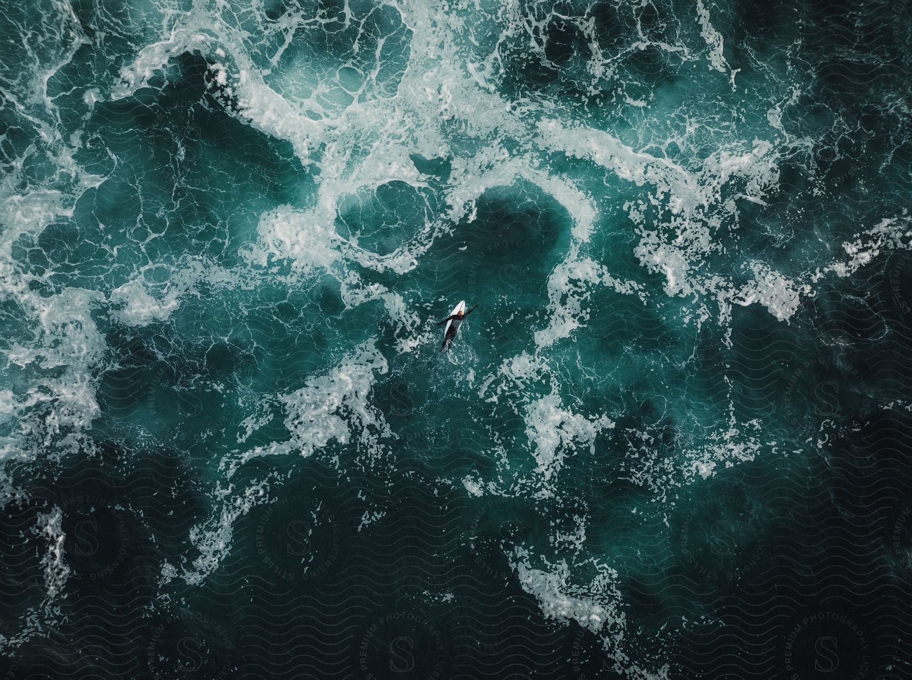 A surfer paddles a surfboard on a turbulent ocean from a topdown aerial perspective