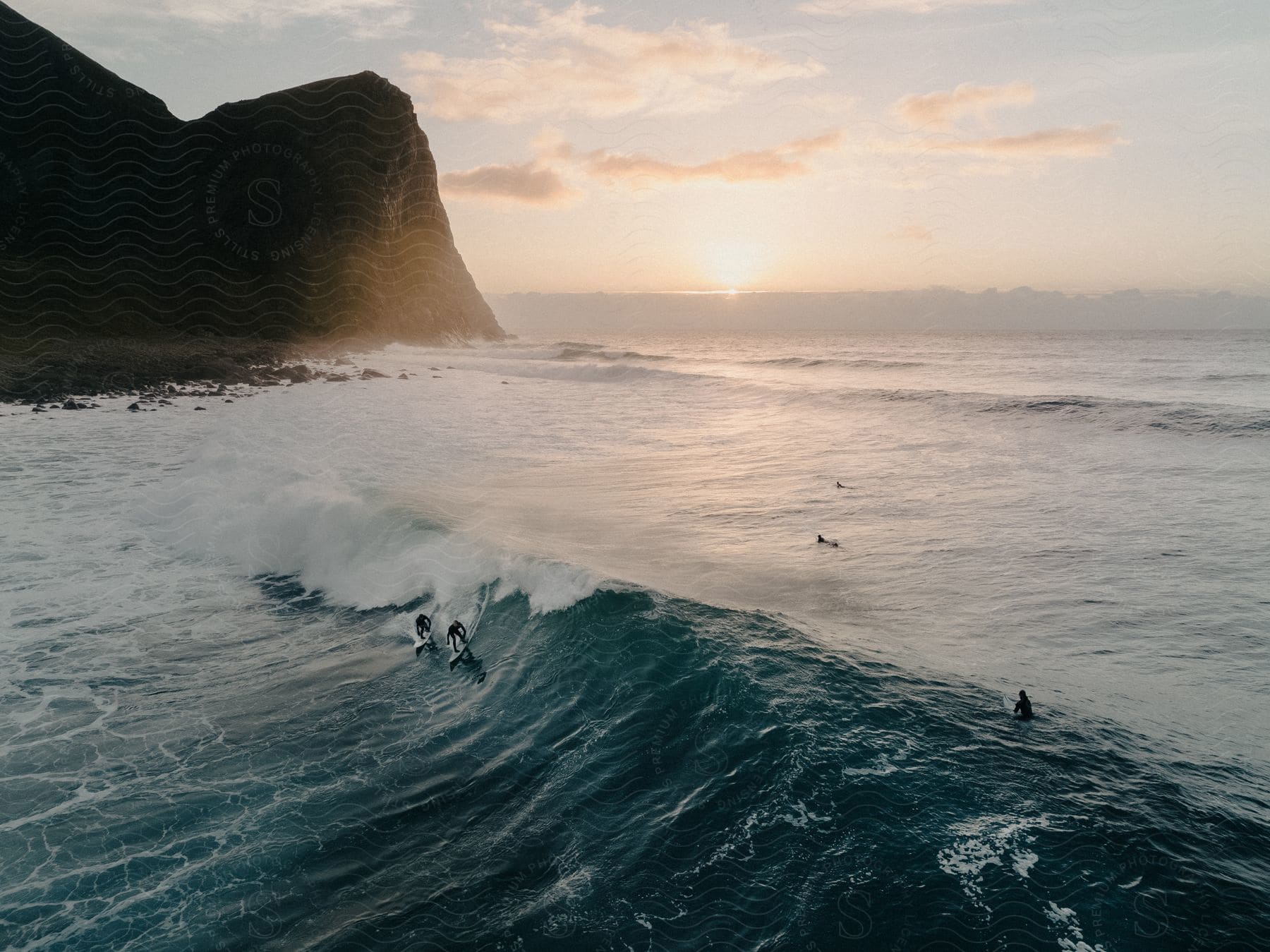 Surfers riding a wave near a promontory as the sun shines on the horizon