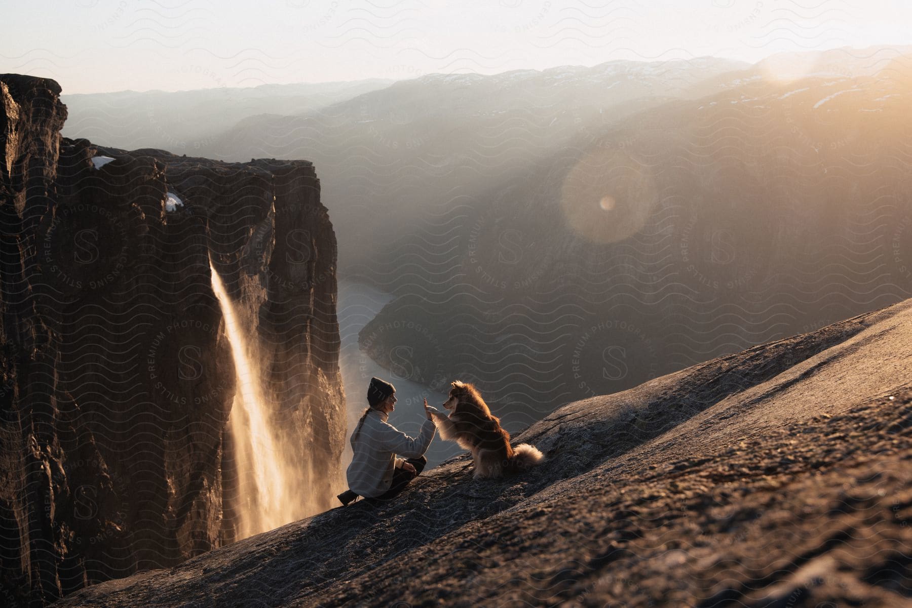 A woman highfives her dog on a cliff near a waterfall