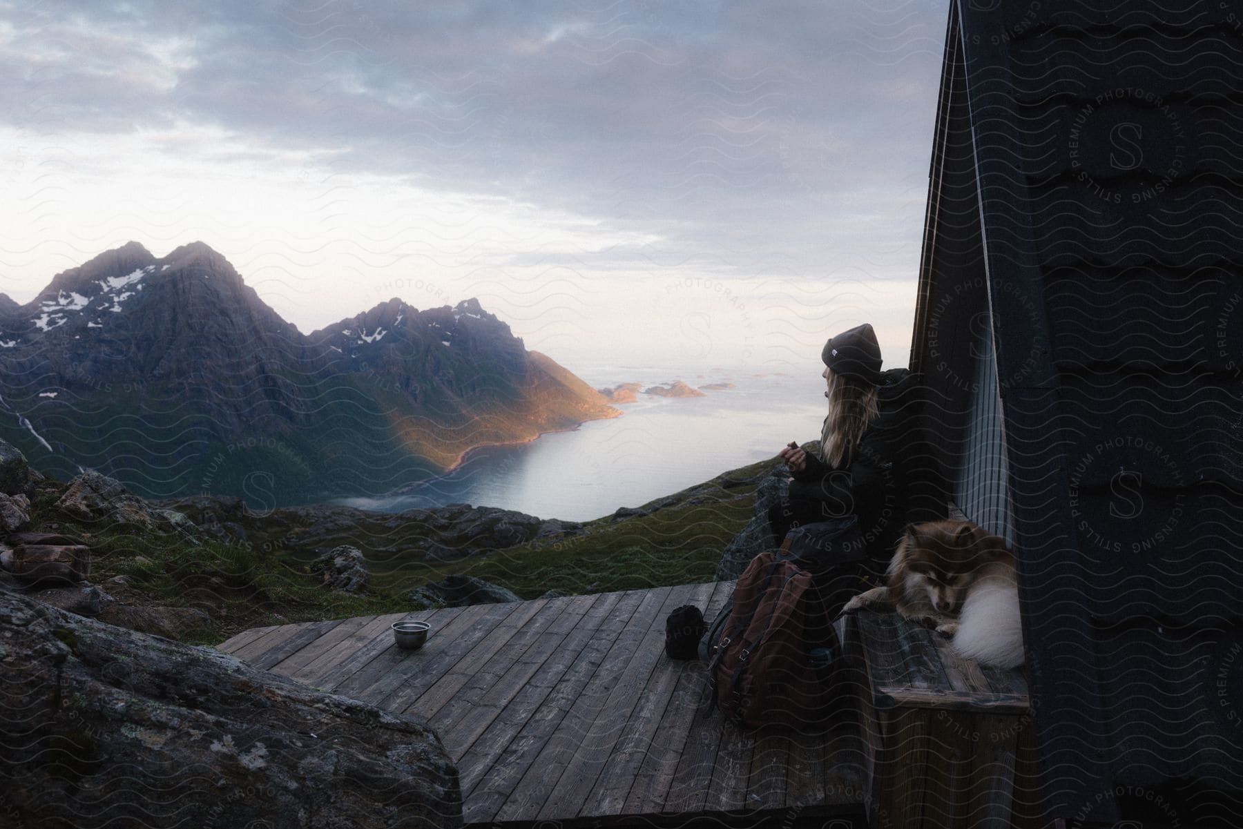 Blonde girl and dog sit outside a bungalow on a mountain with a lake and mountain landscape in the background