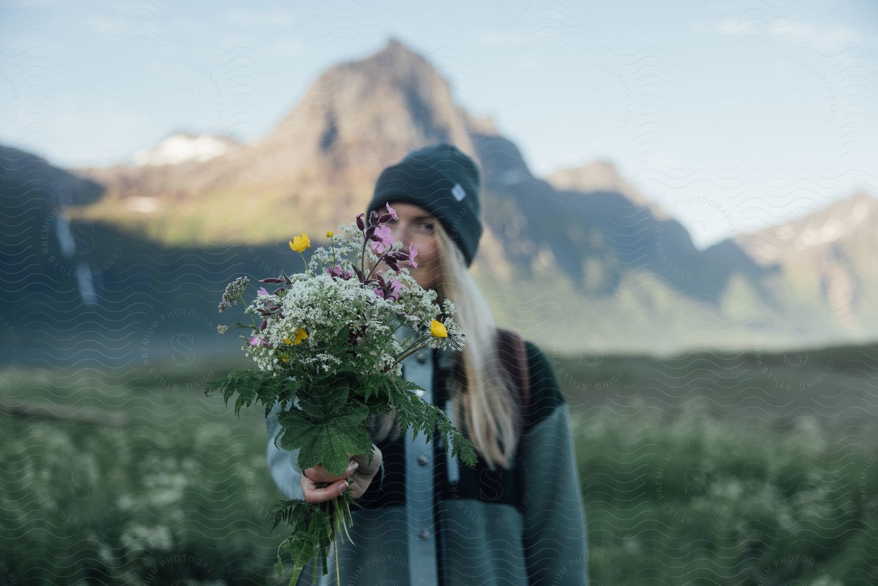 A woman stands on grass in front of a mountain holding flowers in front of her face