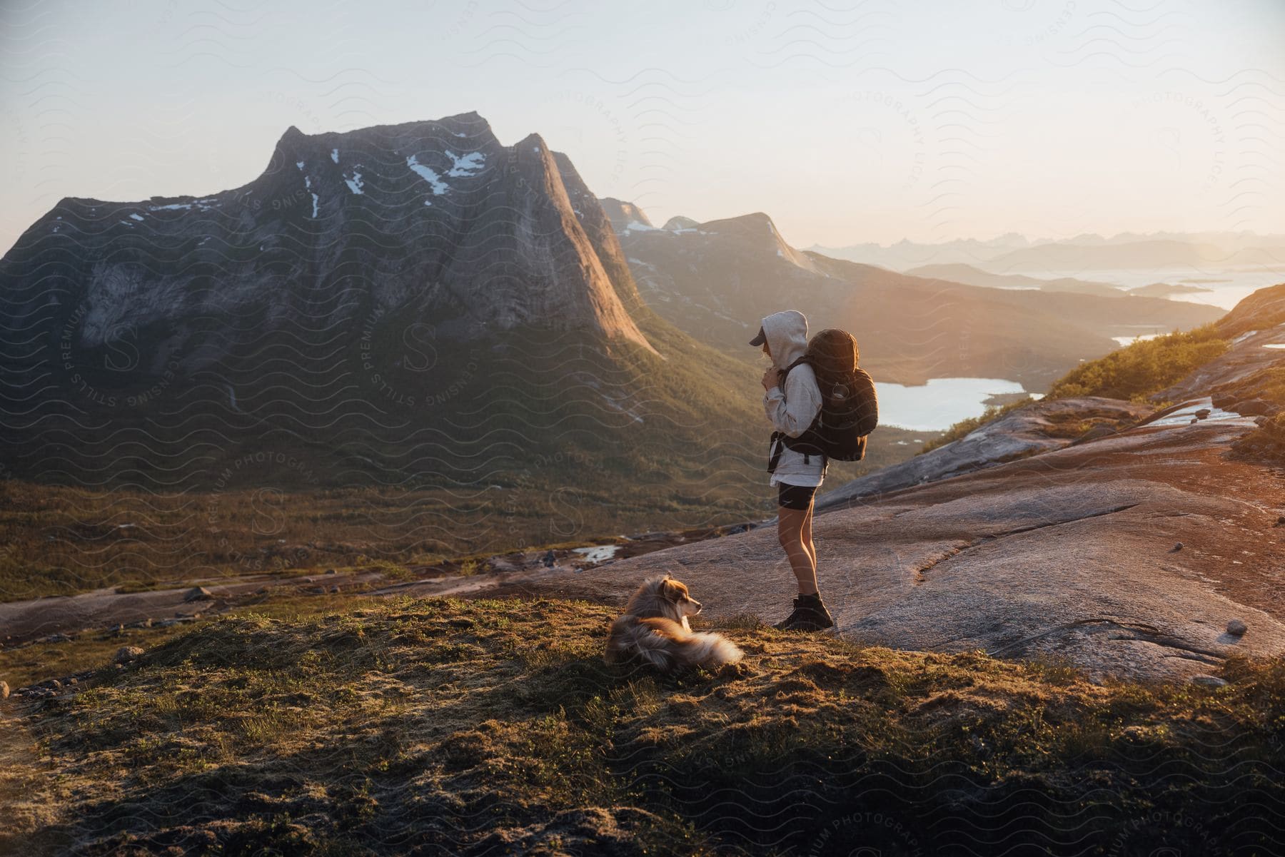 Hiker with backpack stands next to dog near mountains at sunset