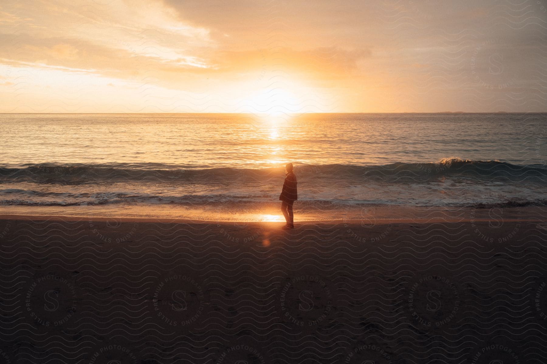 Silhouette of a person standing on a beach as waves roll in and the sun sets on the horizon