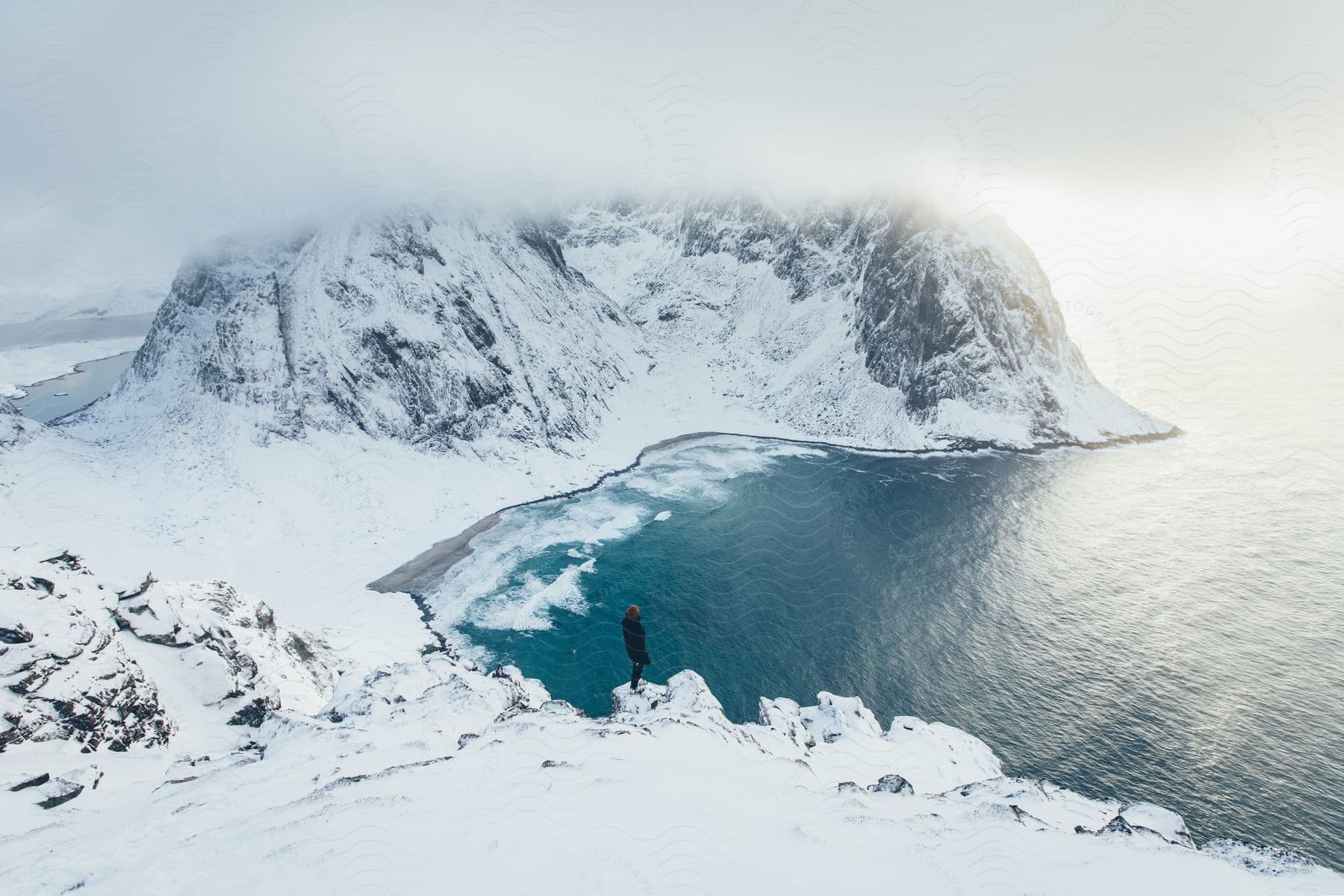 A man stands on a mountain ledge overlooking a snowcovered mountain river valley in norway winter