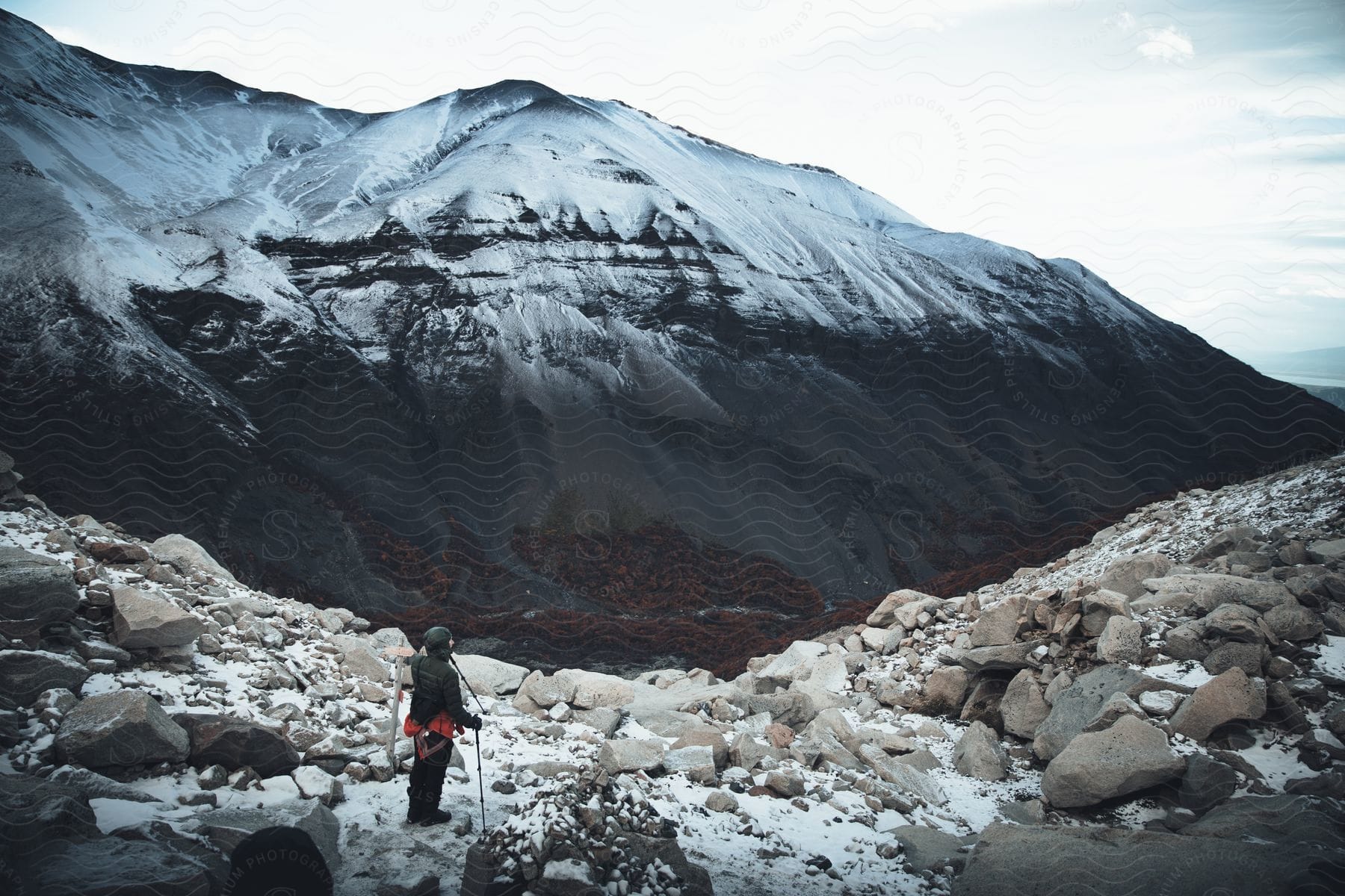 A man stands on a rock covered mountain holding trekking poles looking at a nearby snow covered mountain under a cloudy sky