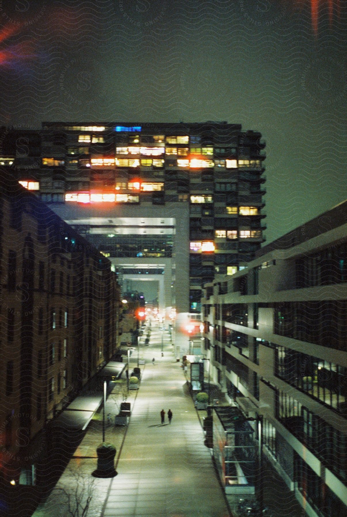 A couple walks down an empty city street at midnight illuminated by street lights with a distant building featuring many lit windows