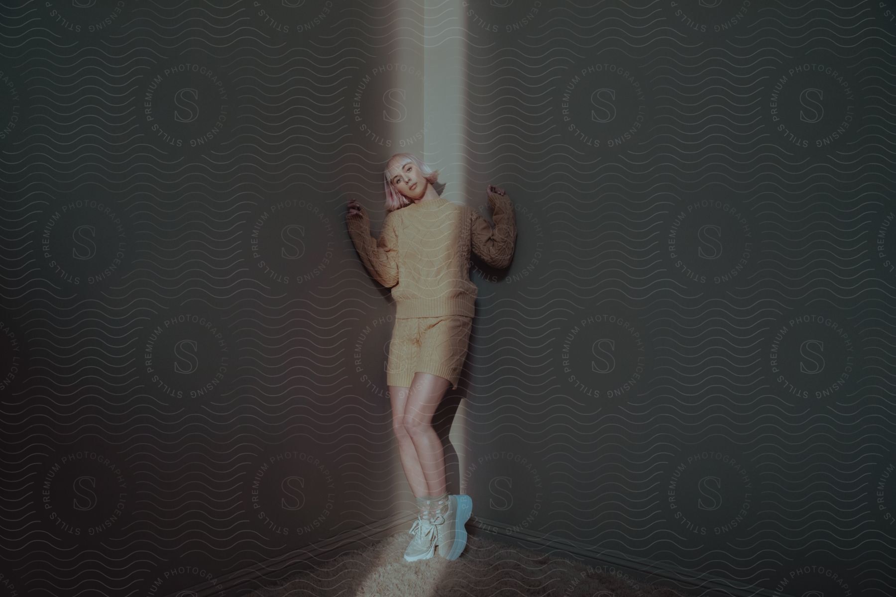 Pink haired woman in woolly clothes standing against a wall in a dark room