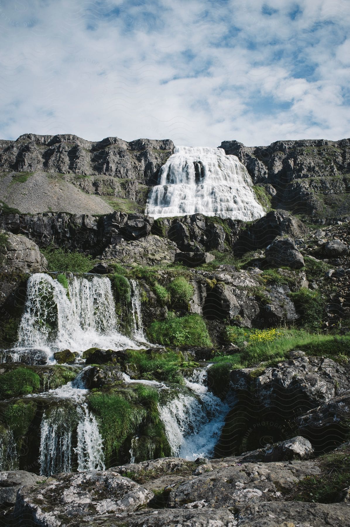 A multitier waterfall with a large white waterfall in the background and a green field below it with a few yellow flowers