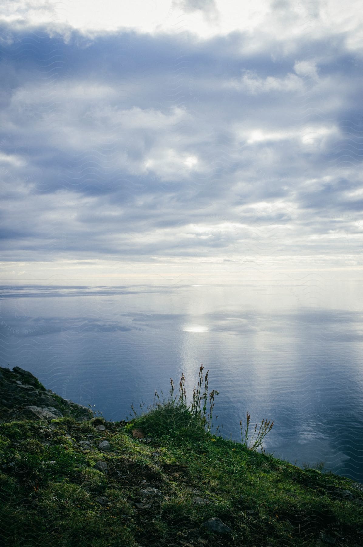 Sun shines through rain reflecting on sea beyond mossy coastal hill