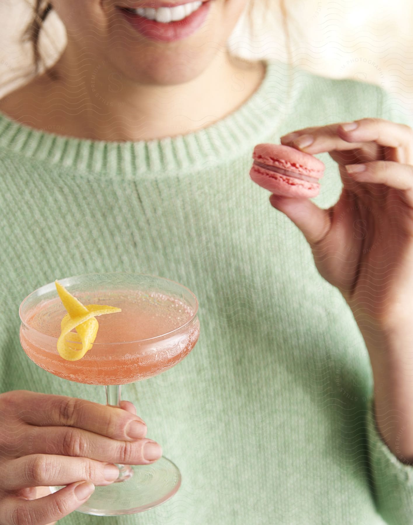 A woman in a green sweater holds a pink drink and a pink cookie