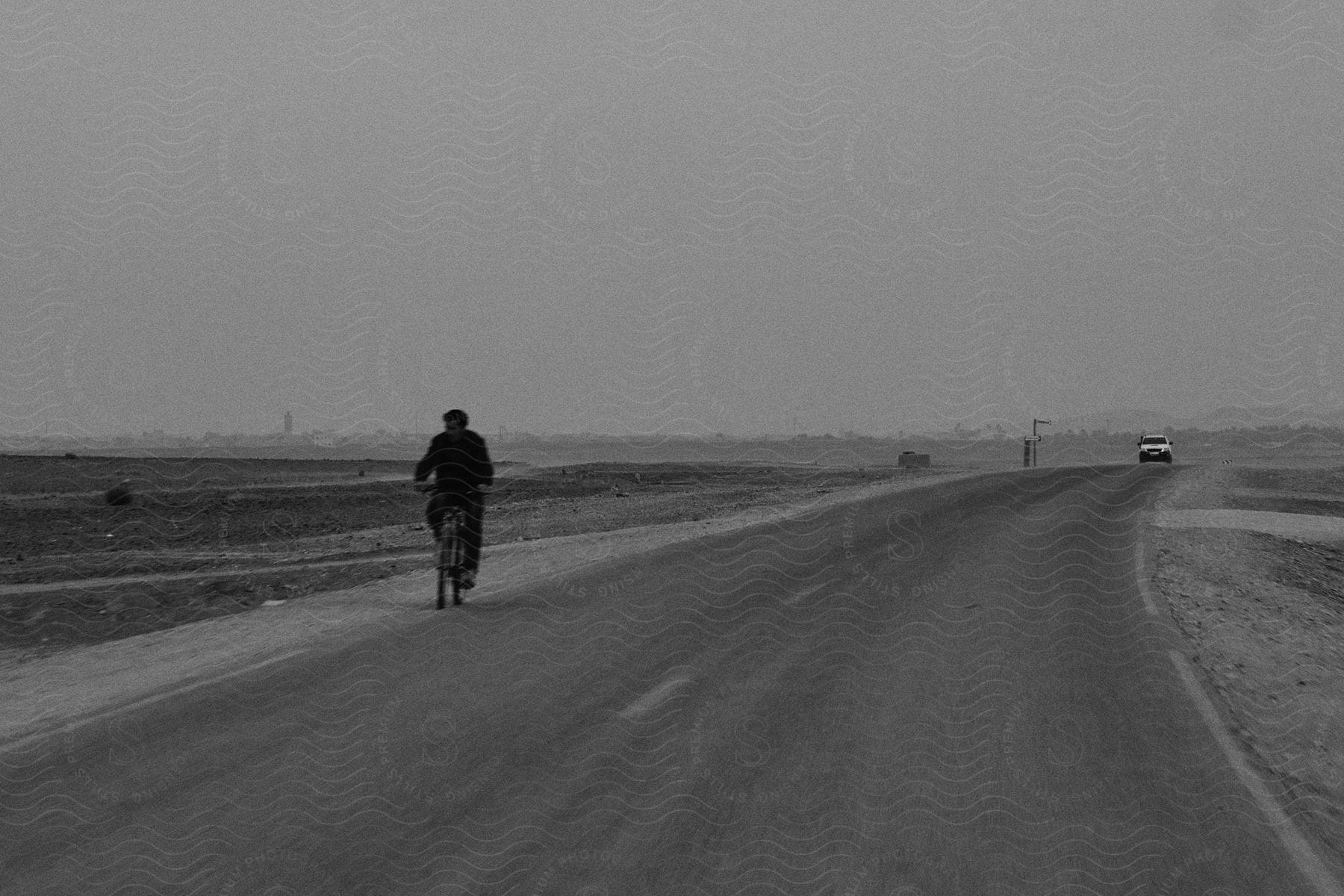 A man on a bicycle rides along a paved highway road with a car approaching behind him