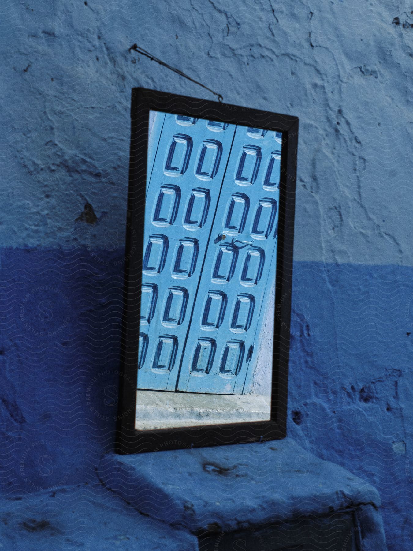 A blue door reflected in a rectangular mirror hanging on a blue cement wall