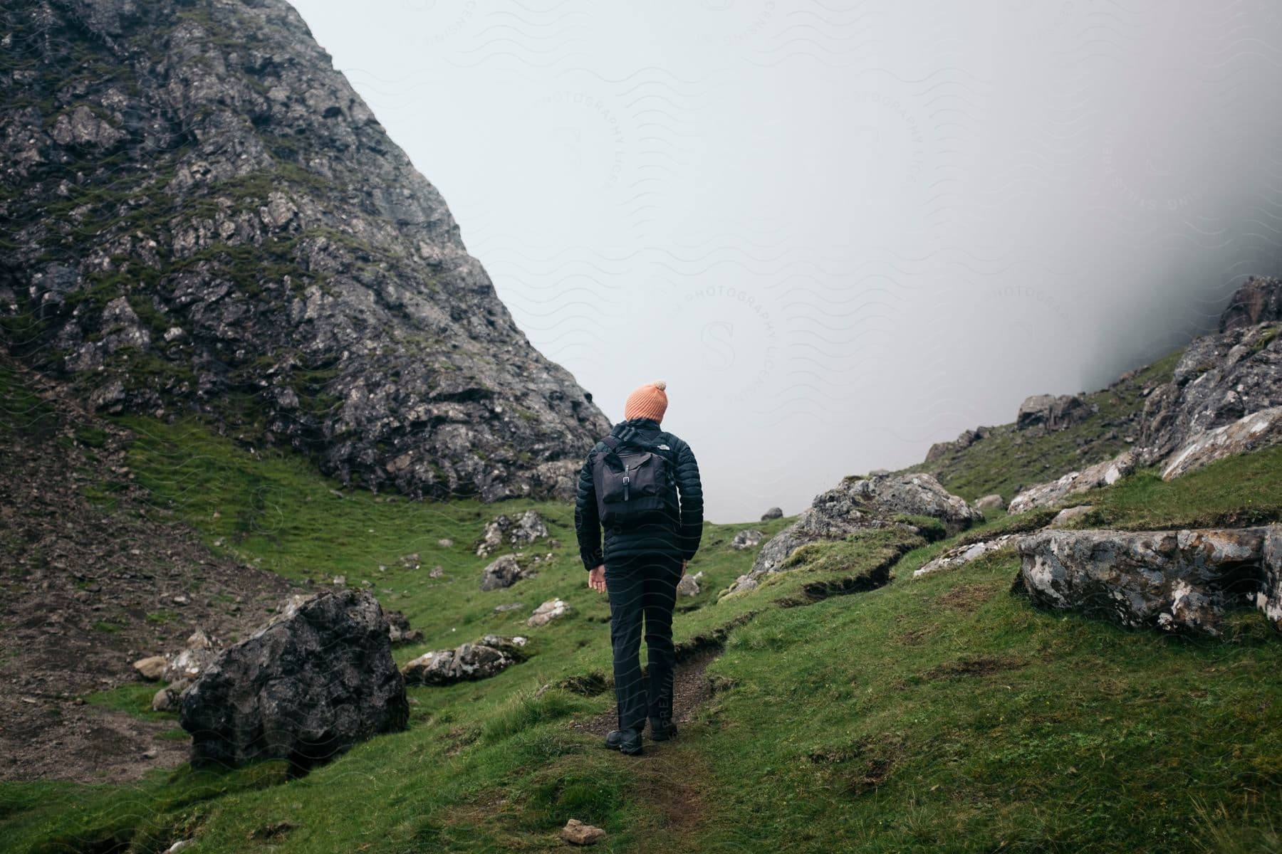 A man walking on a trail towards foggy mountains with a backpack and jacket on