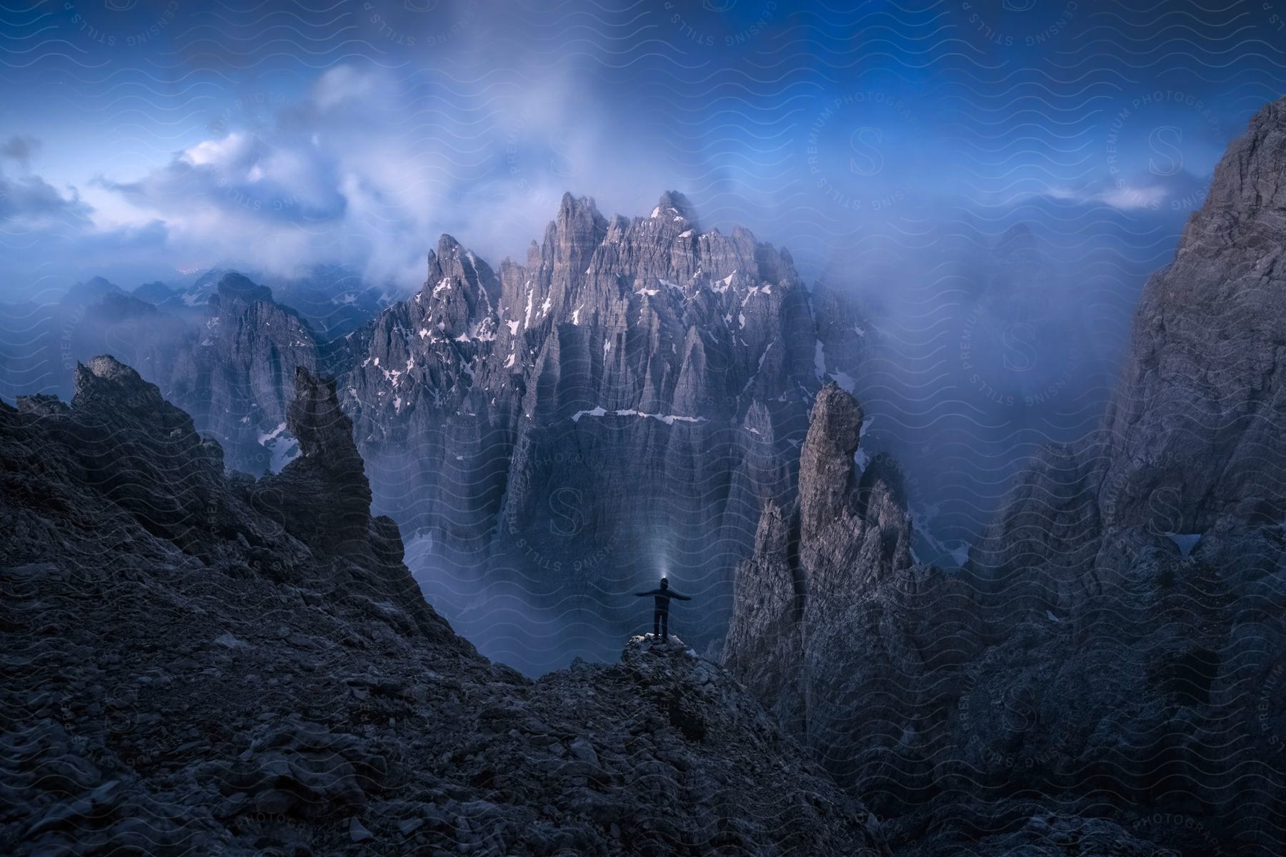 A man standing on a mountain ledge with arms spread facing a mountain ridge at dusk