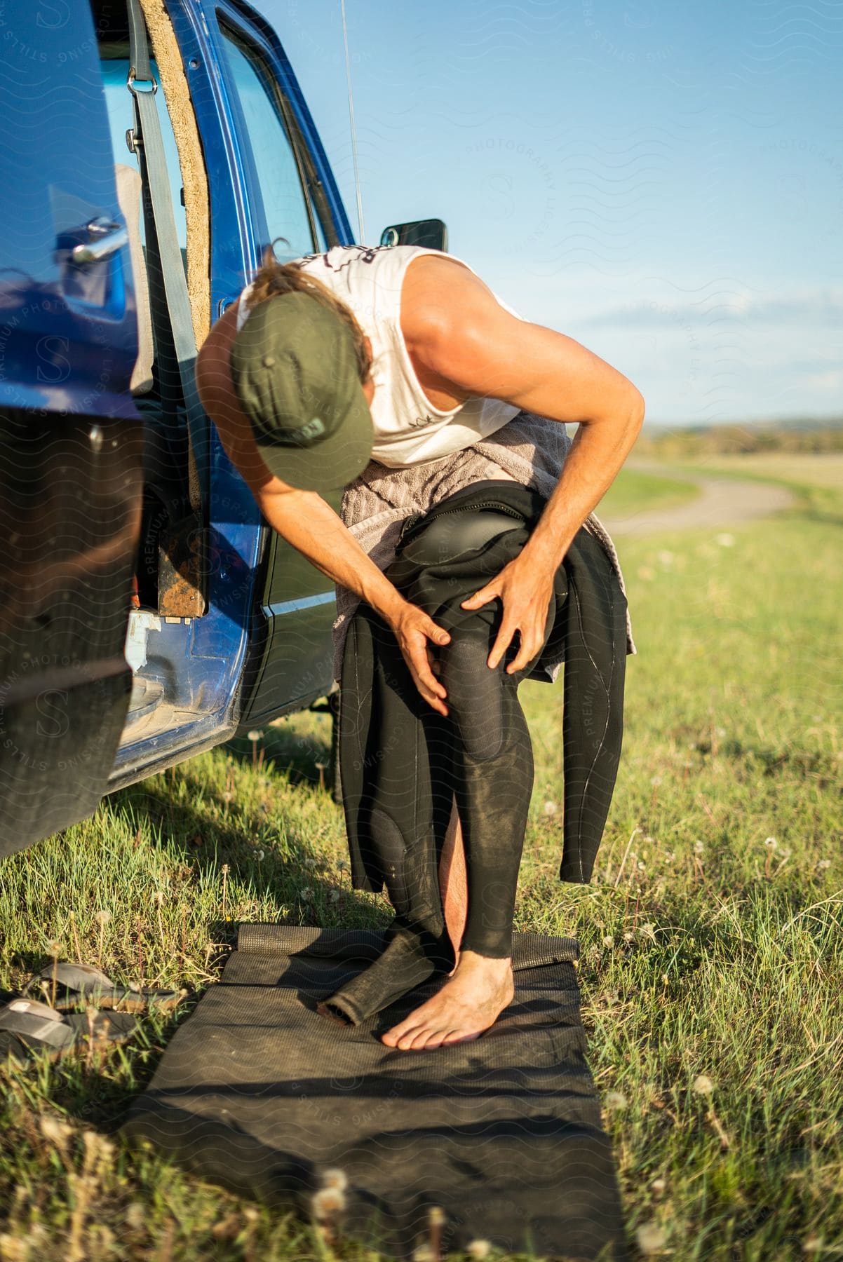 Man wearing a cap and white tank top puts on a scuba diving wetsuit next to a van in plain fields