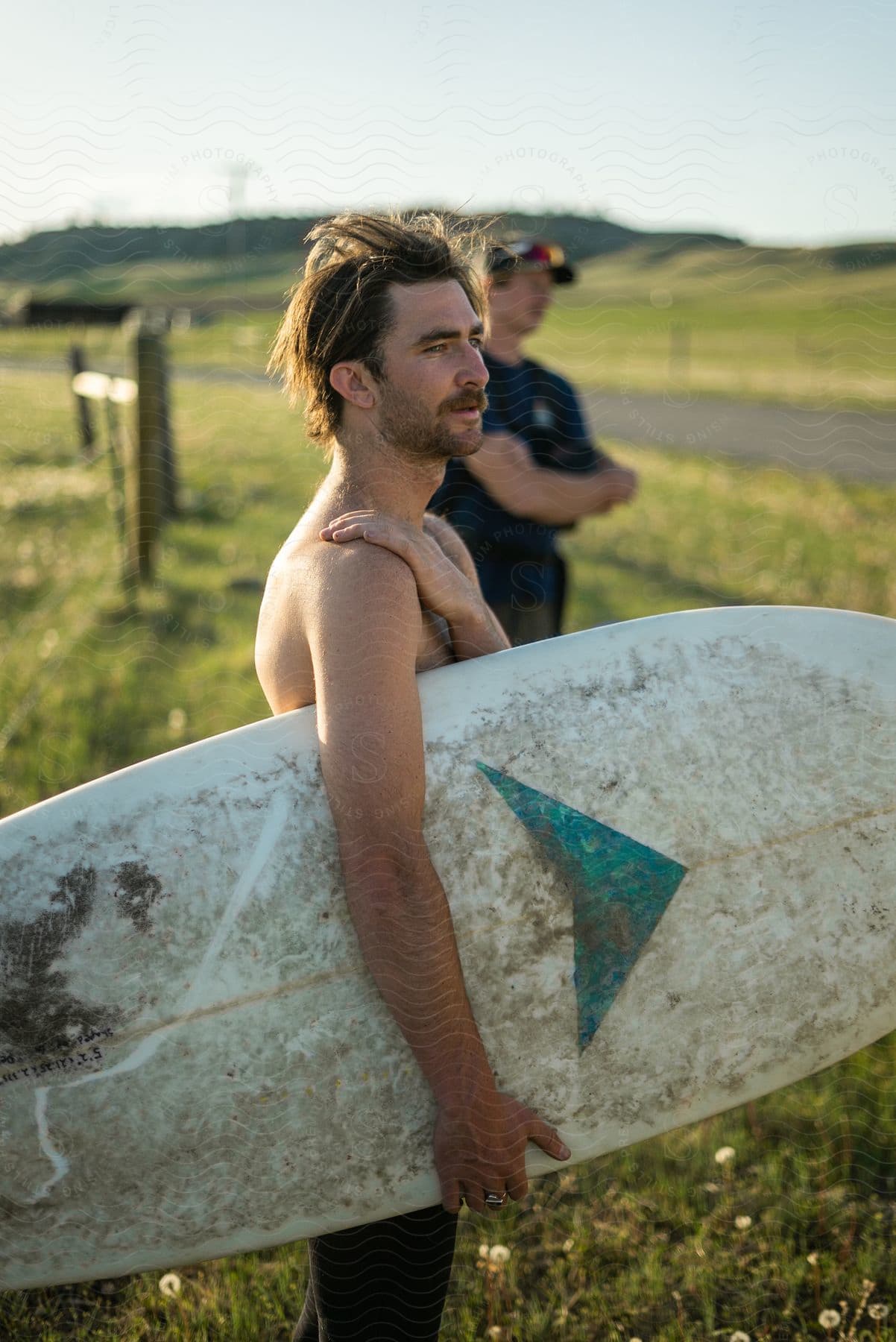 A man stands in a grassy field holding a white surfboard with another man out of focus nearby